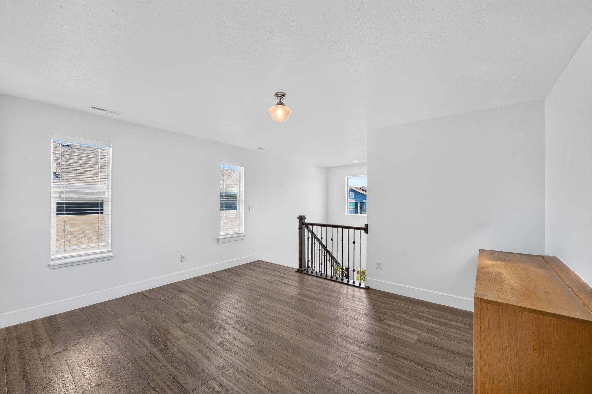 Upper-level landing featuring wood-finish flooring, white walls, two windows with blinds, a black metal staircase railing, and a ceiling-mounted light fixture