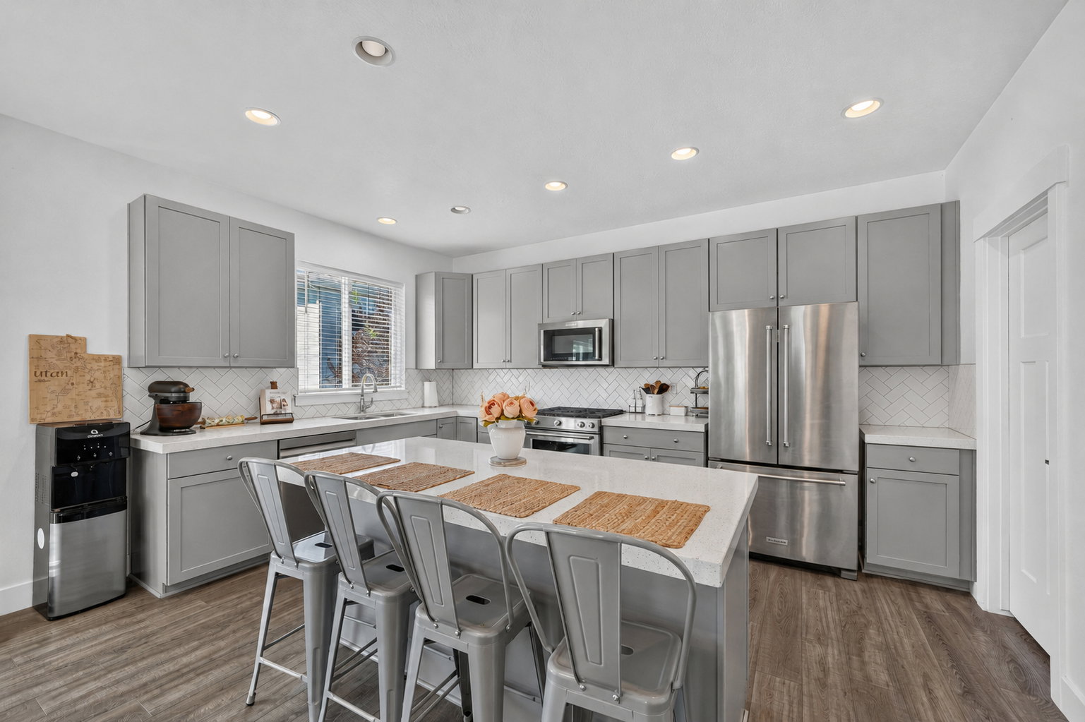 Modern kitchen featuring a central island with seating for four, grey shaker-style cabinetry, stainless steel appliances, a white herringbone tile backsplash, and wood-finish flooring