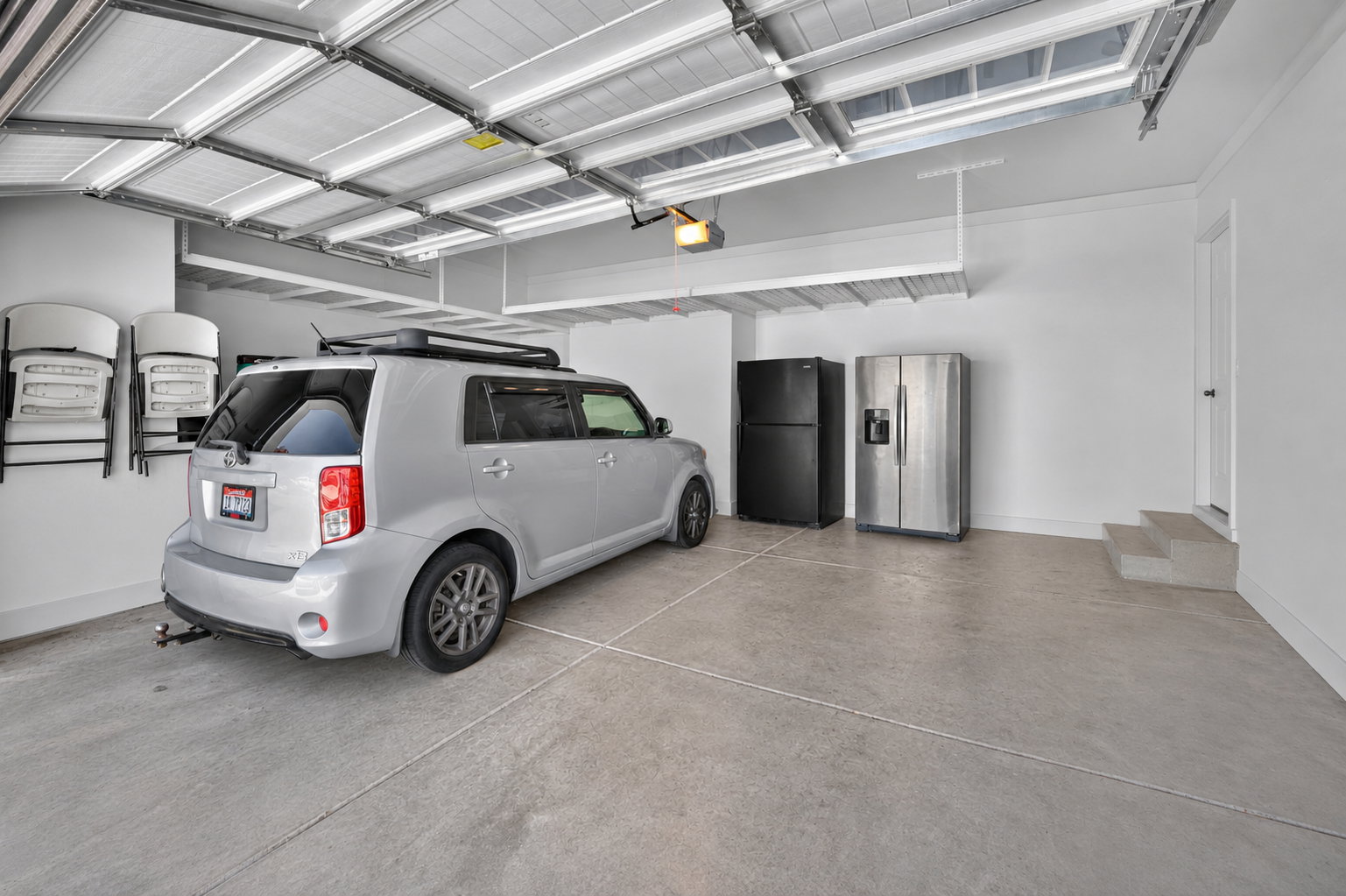 Spacious garage featuring a finished concrete floor, overhead storage racks, a white paneled garage door, crisp white walls, and built-in steps leading to an interior door