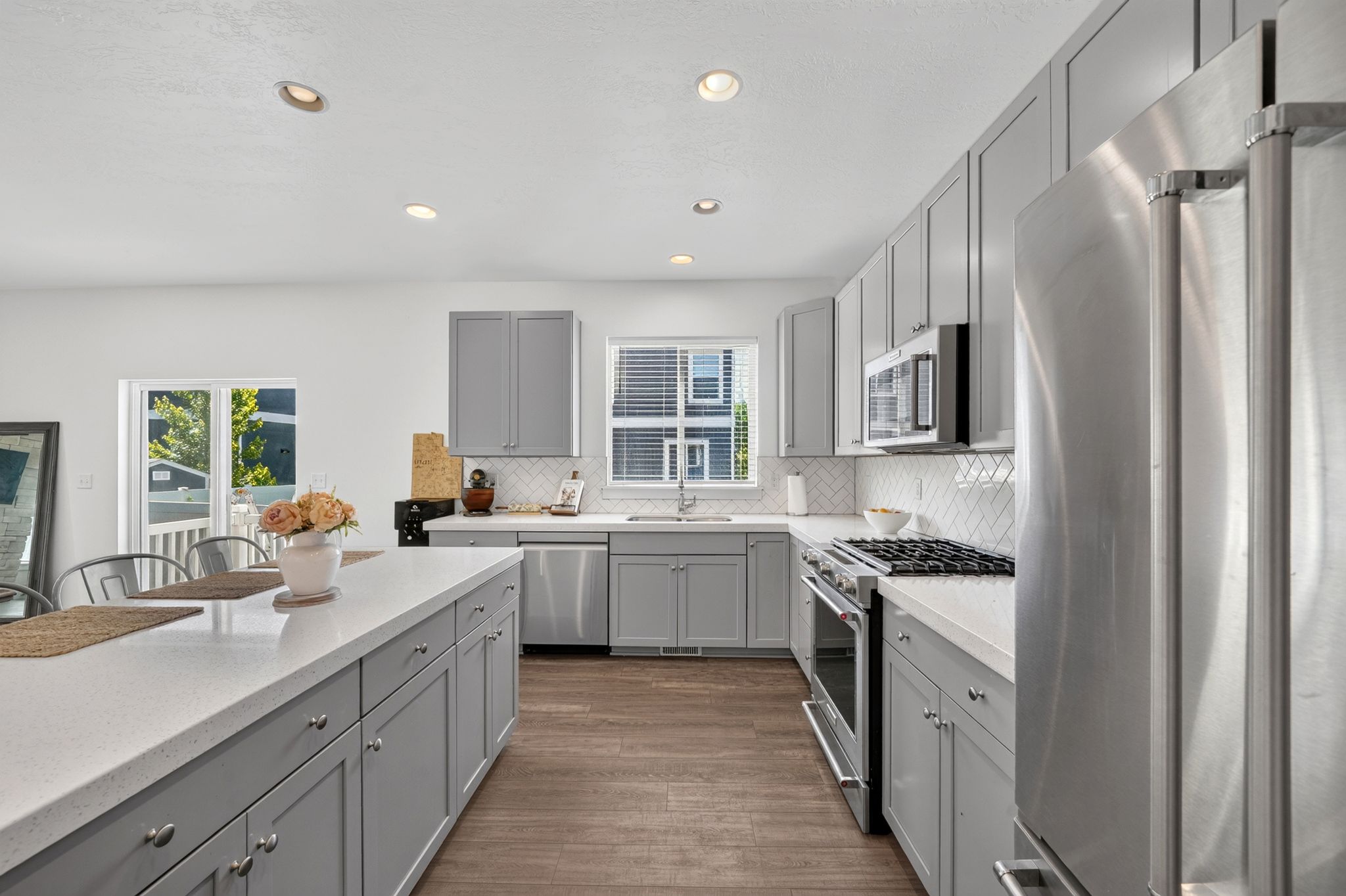 Spacious kitchen featuring an extended island with a light-toned countertop, gray shaker-style cabinetry, a herringbone tile backsplash, recessed lighting, and stainless steel appliances including a gas range