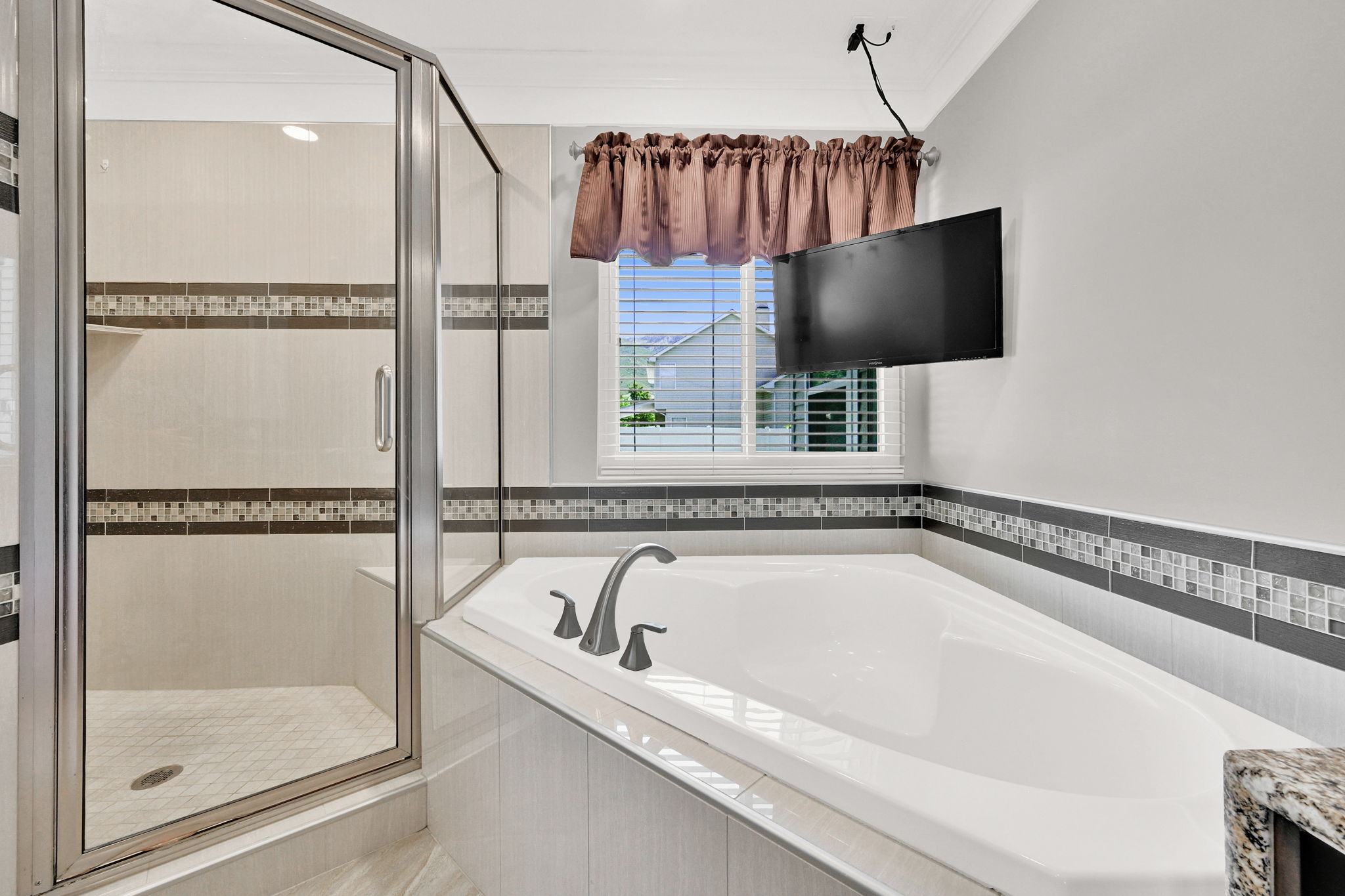 Primary ensuite with built-in soaking tub with a tiled surround, complemented by a glass-enclosed shower featuring a tiled bench and mosaic accent banding and a television