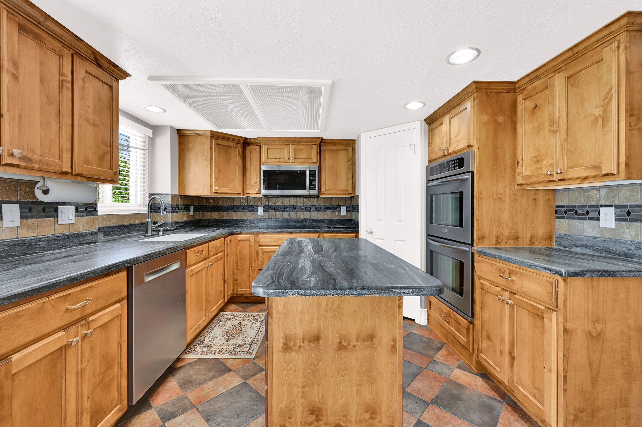 Kitchen featuring natural wood cabinetry, stone countertops, a mosaic tile backsplash, stainless steel appliances, and a central movable island