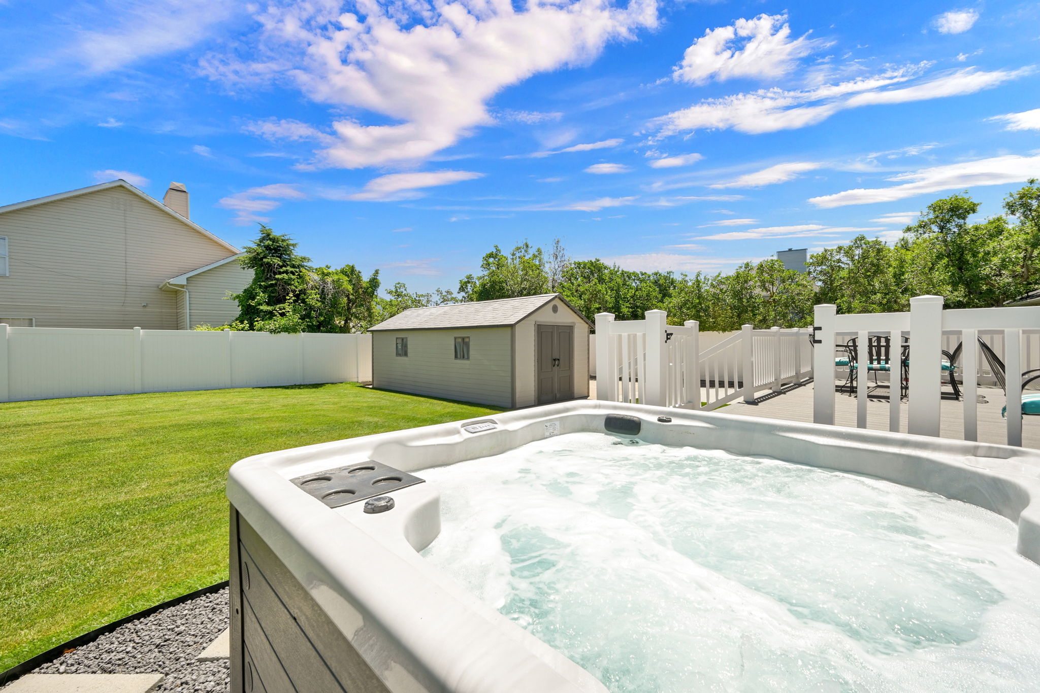 Outdoor hot tub with textured gray siding and stone surround, accompanied by a spacious green lawn, a white privacy fence, an outdoor storage sheds, and a composite deck with white railings