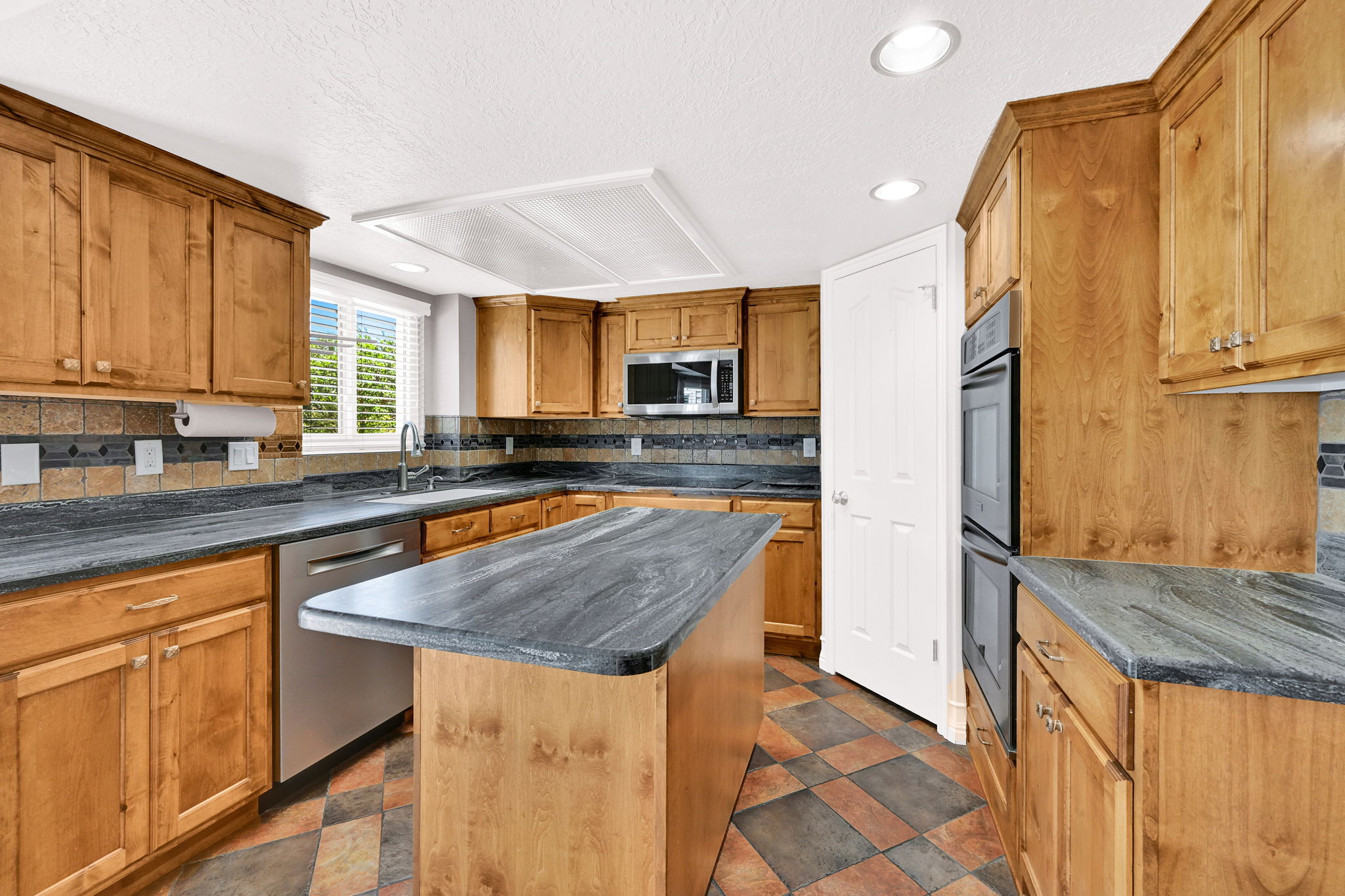 Kitchen featuring natural wood cabinetry, dark patterned countertops, stainless steel appliances, and a tiled backsplash