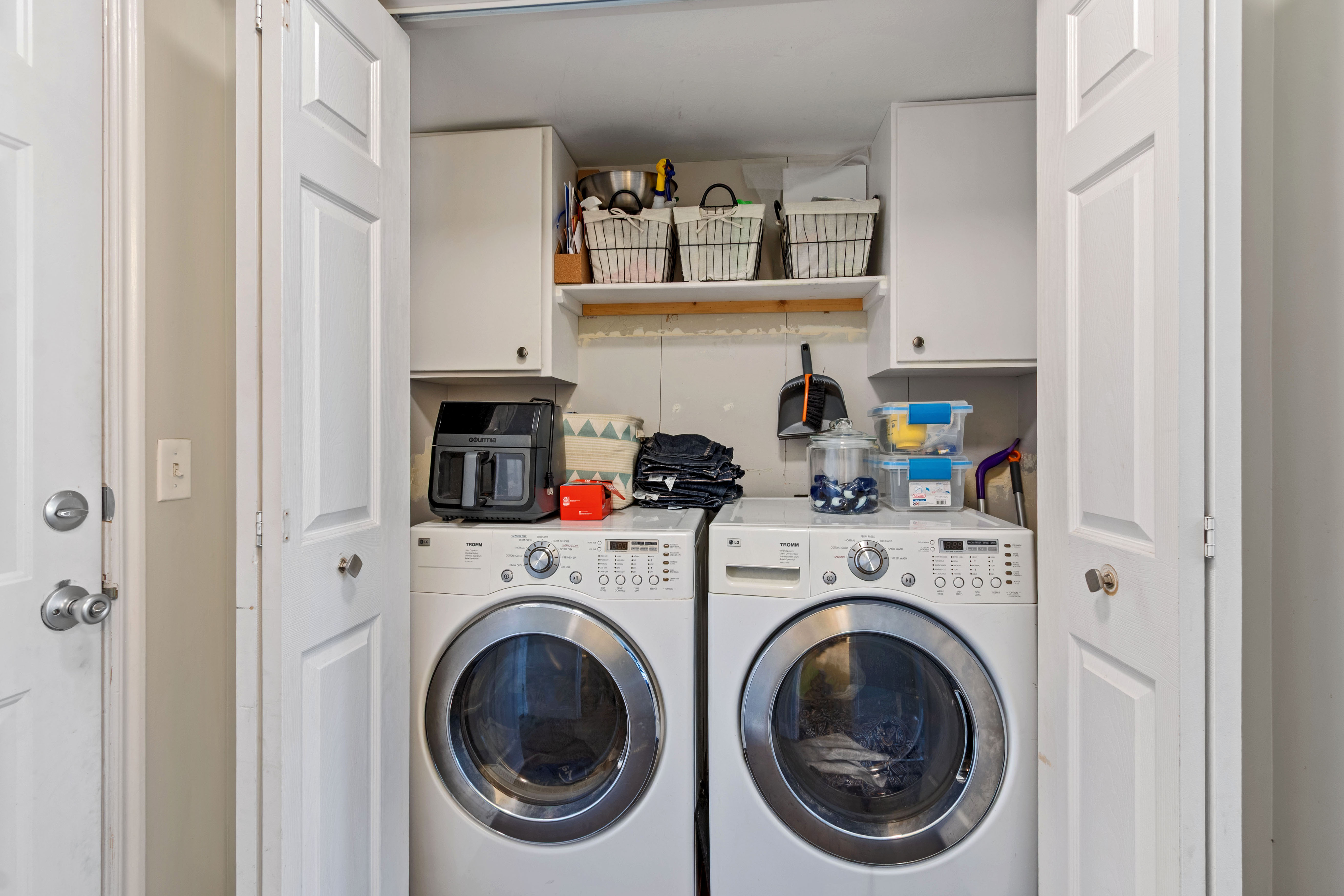 Dedicated laundry area featuring white bi-fold doors, upper cabinetry, an open shelf, and white front-load appliances