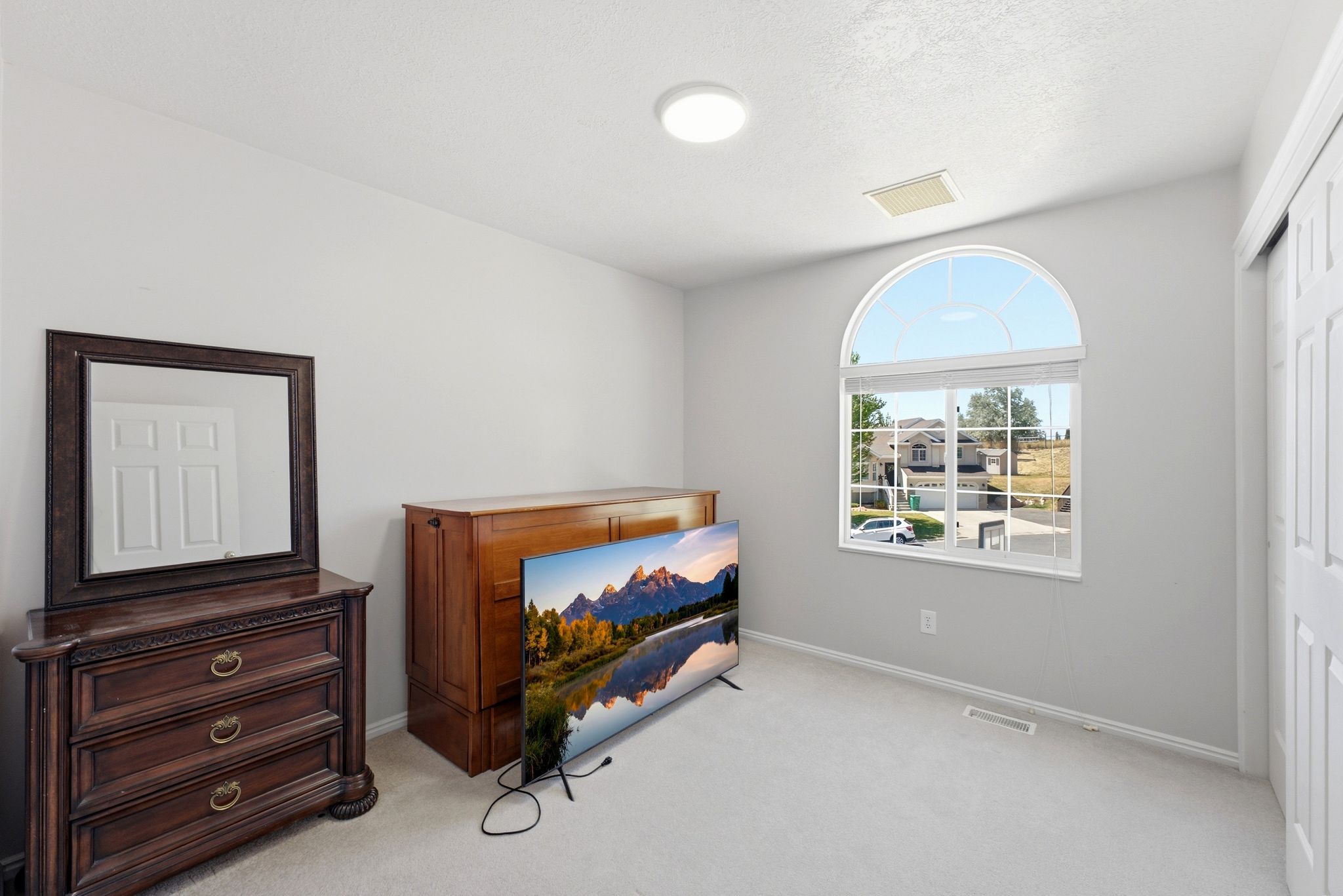 Bright room featuring a large arched window with white trim, soft gray walls, light-colored carpeting, and a built-in closet with bi-fold doors