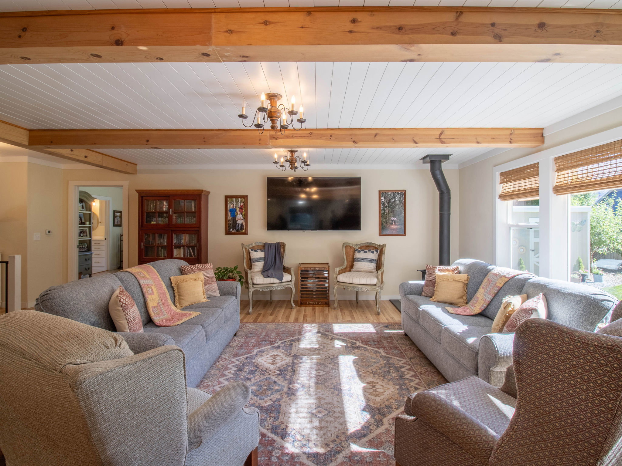 Spacious common area featuring wood-finish flooring, exposed ceiling beams, a wood-burning stove, large windows with natural light, and a decorative chandelier