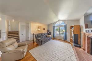 Living area featuring vaulted ceilings, wood-finish flooring, a prominent arched window, a brick fireplace with a wood mantel, and built-in shelving