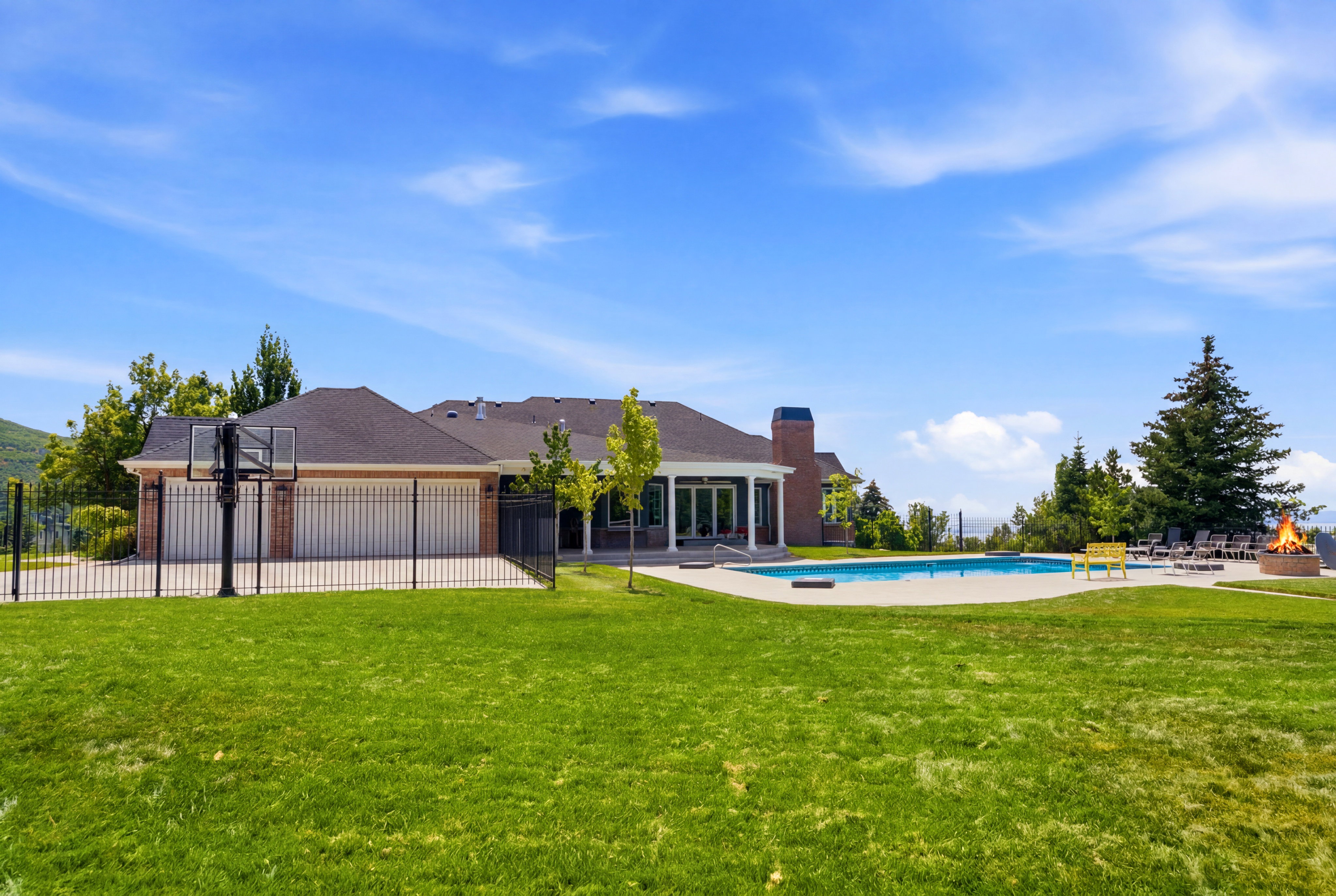 Expansive green lawn featuring an inground swimming pool with surrounding patio, a basketball court with black fencing, and a built-in fire pit
