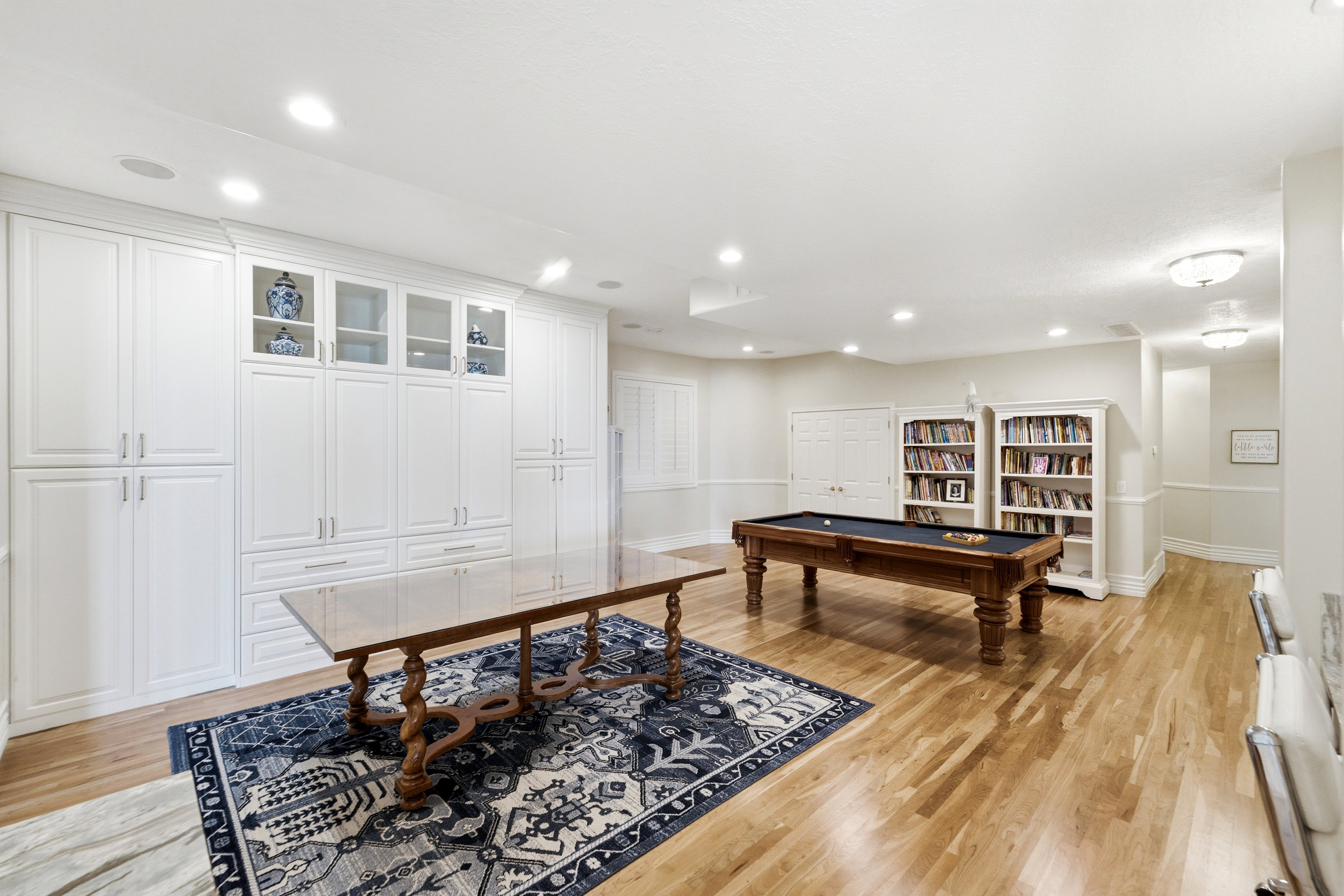 Spacious recreation room featuring extensive built-in white cabinetry with glass-front display uppers, hardwood flooring, recessed lighting, and a wide hallway with decorative wall molding