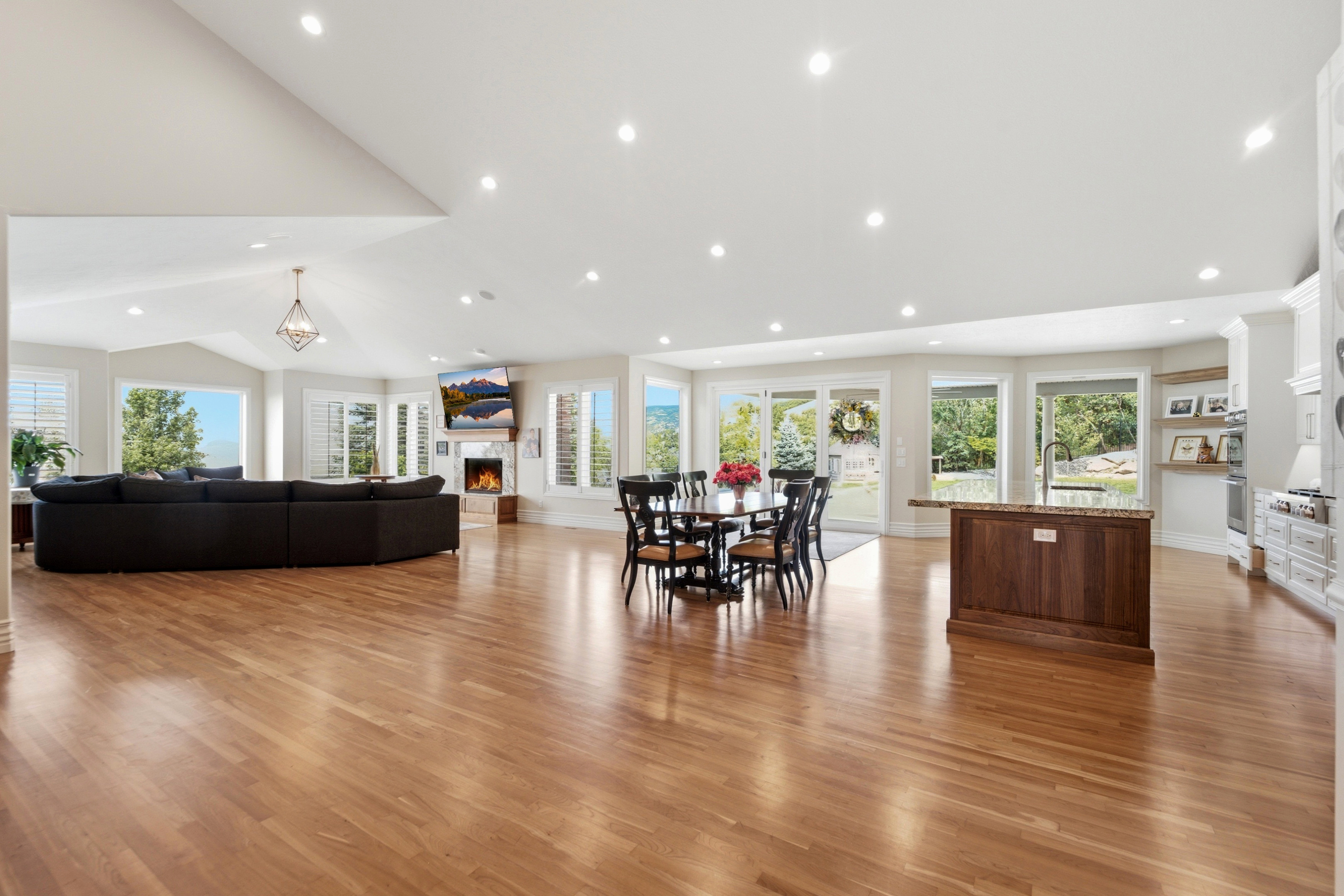 Expansive open-concept living area featuring wood-finish flooring, abundant recessed lighting, multiple large windows with plantation shutters, a central fireplace with stone surround, and an integrated kitchen island with a granite countertop