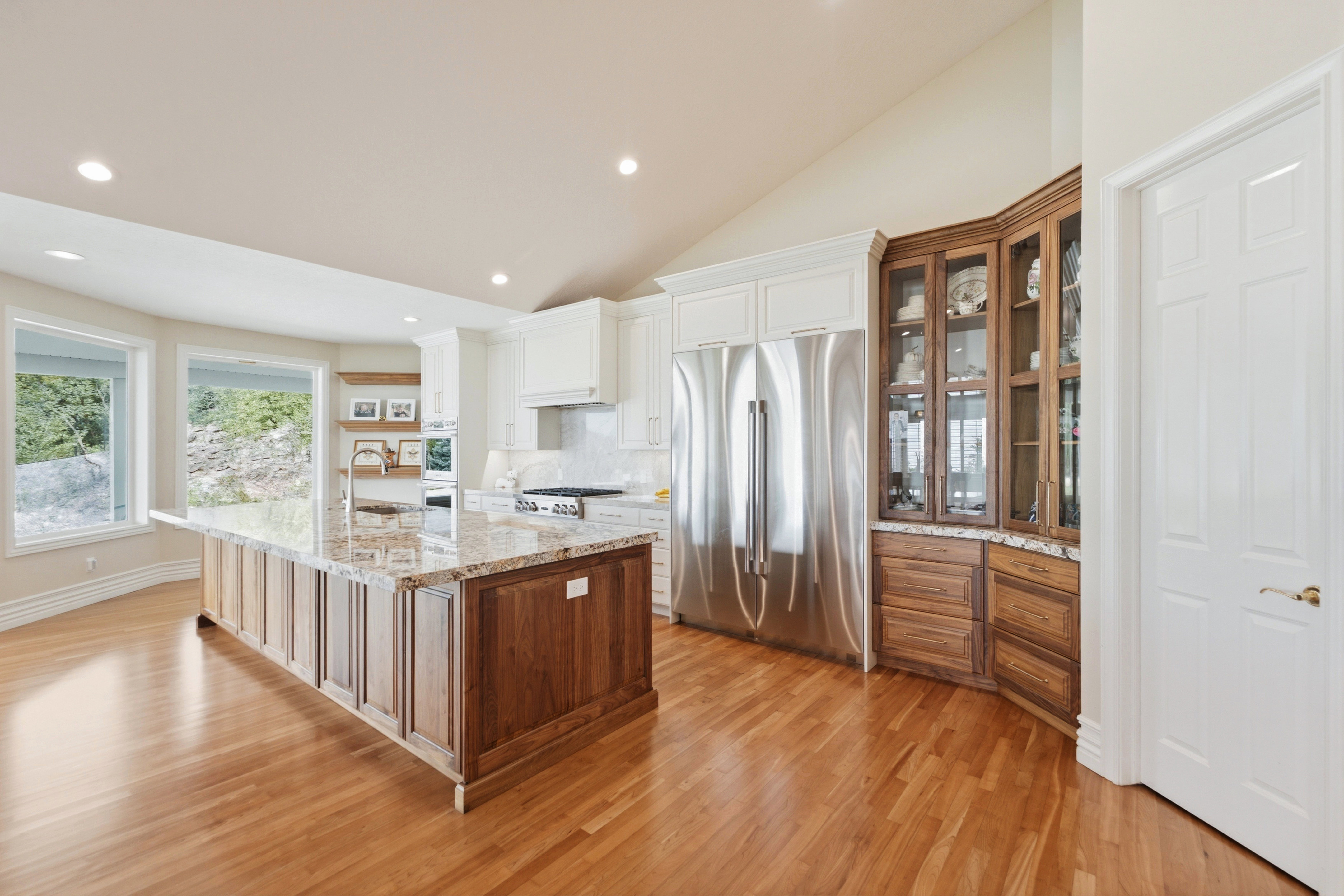Kitchen featuring a large central island with a granite countertop, stainless steel appliances, light wood-finish flooring, white upper cabinetry, and a vaulted ceiling