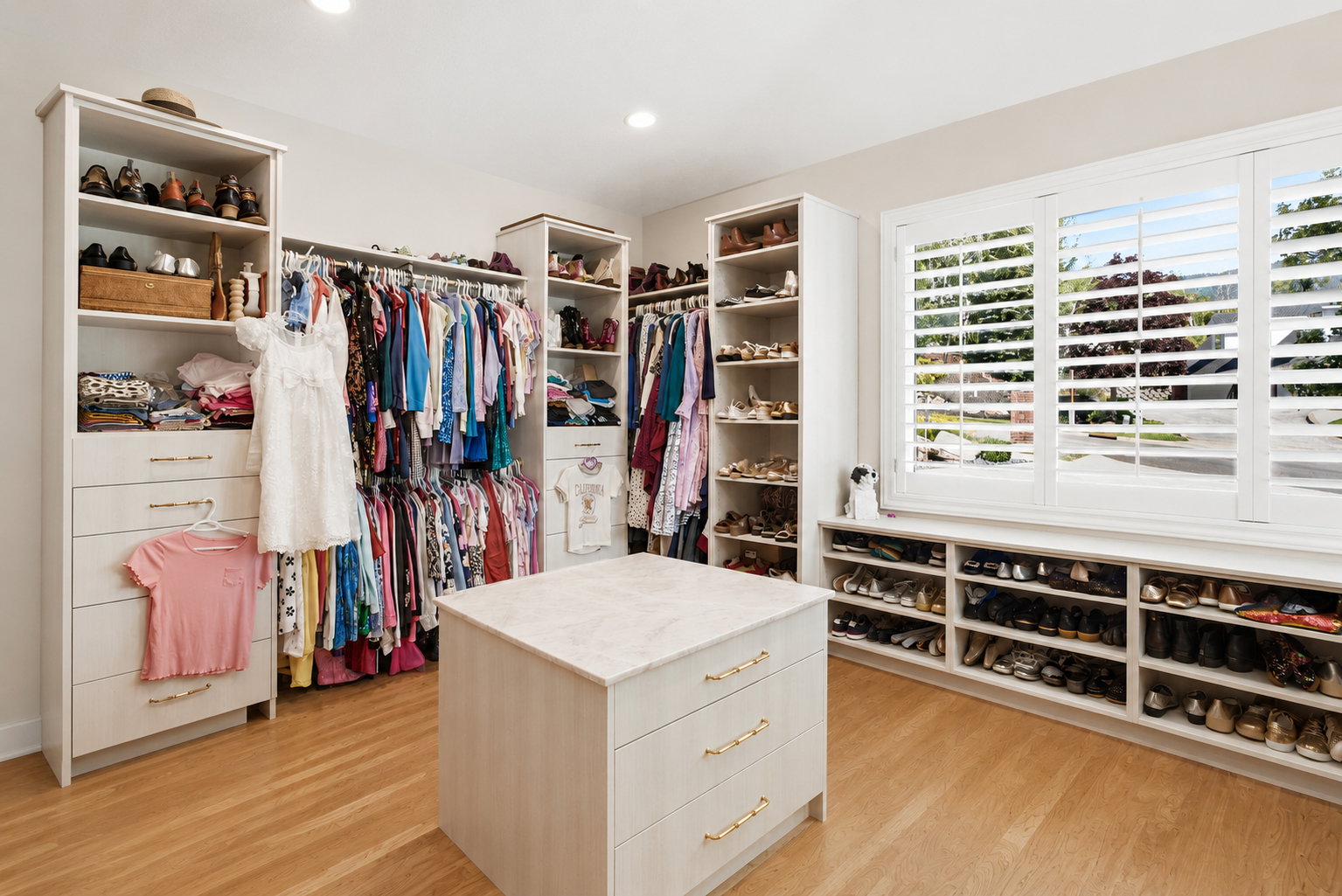 Custom closet system featuring wood-finish flooring, built-in shelving, an organizational island with stone-finish countertop, window-mounted plantation shutters, and recessed ceiling lighting