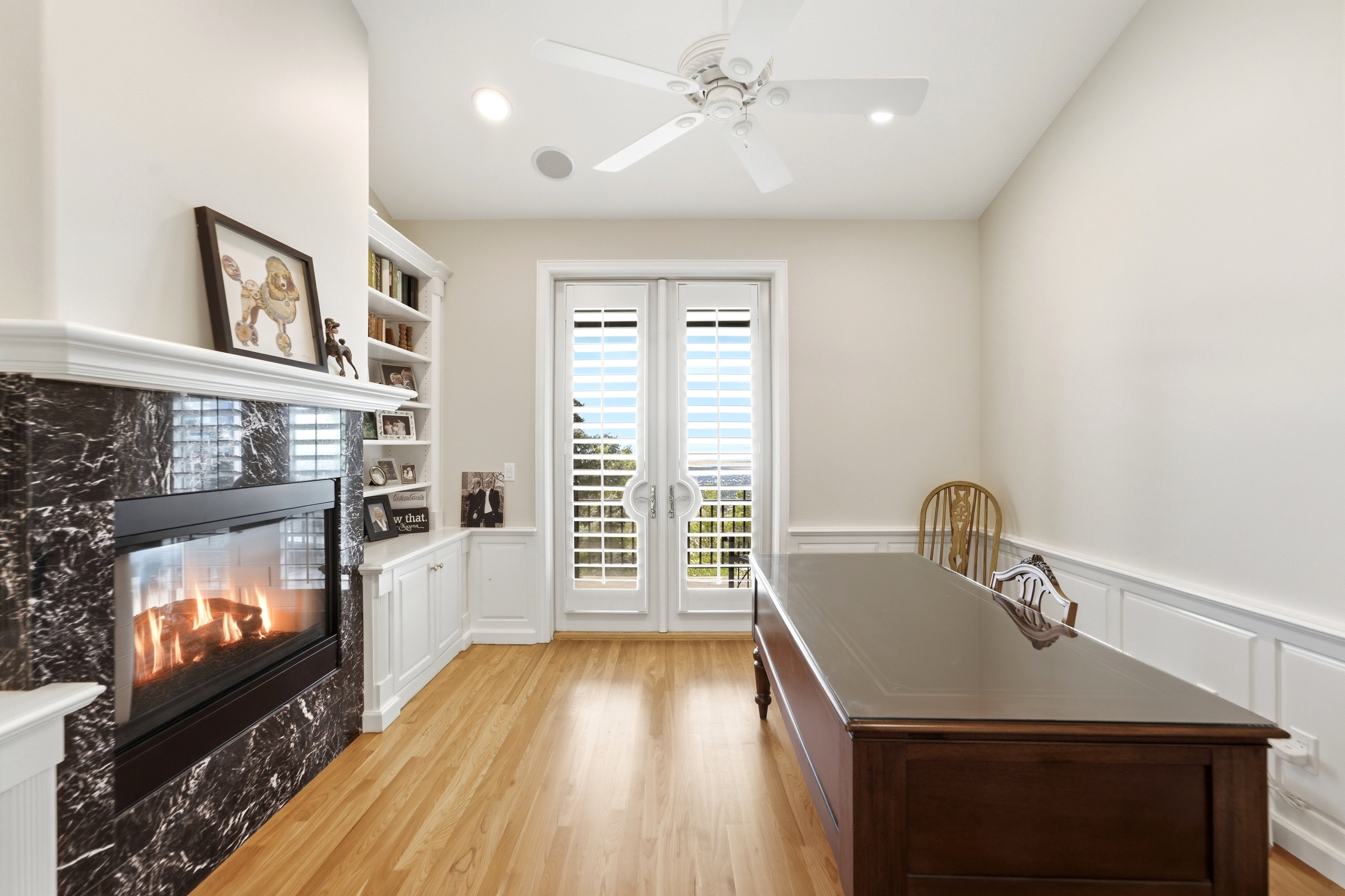Room featuring light wood-finish flooring, a contemporary fireplace with a dark stone surround and white mantel, built-in shelving, white paneled French doors with plantation shutters, and a ceiling fan with integrated lighting