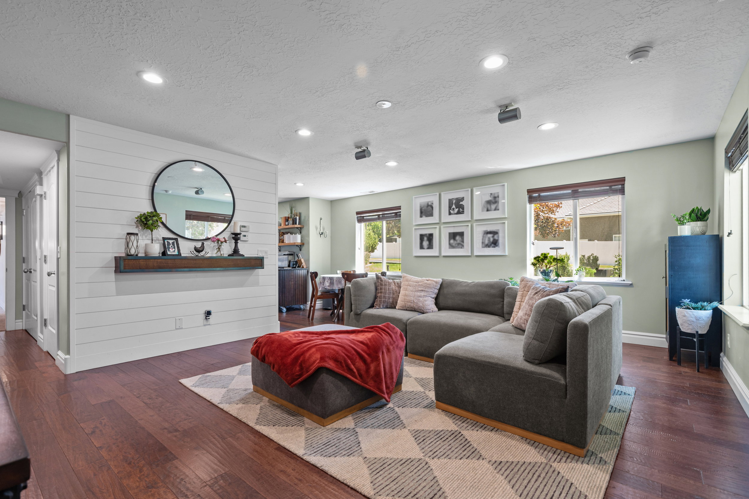 Open-concept living area featuring wood-finish flooring, a prominent shiplap accent wall with a floating shelf, recessed lighting, and multiple windows with blinds