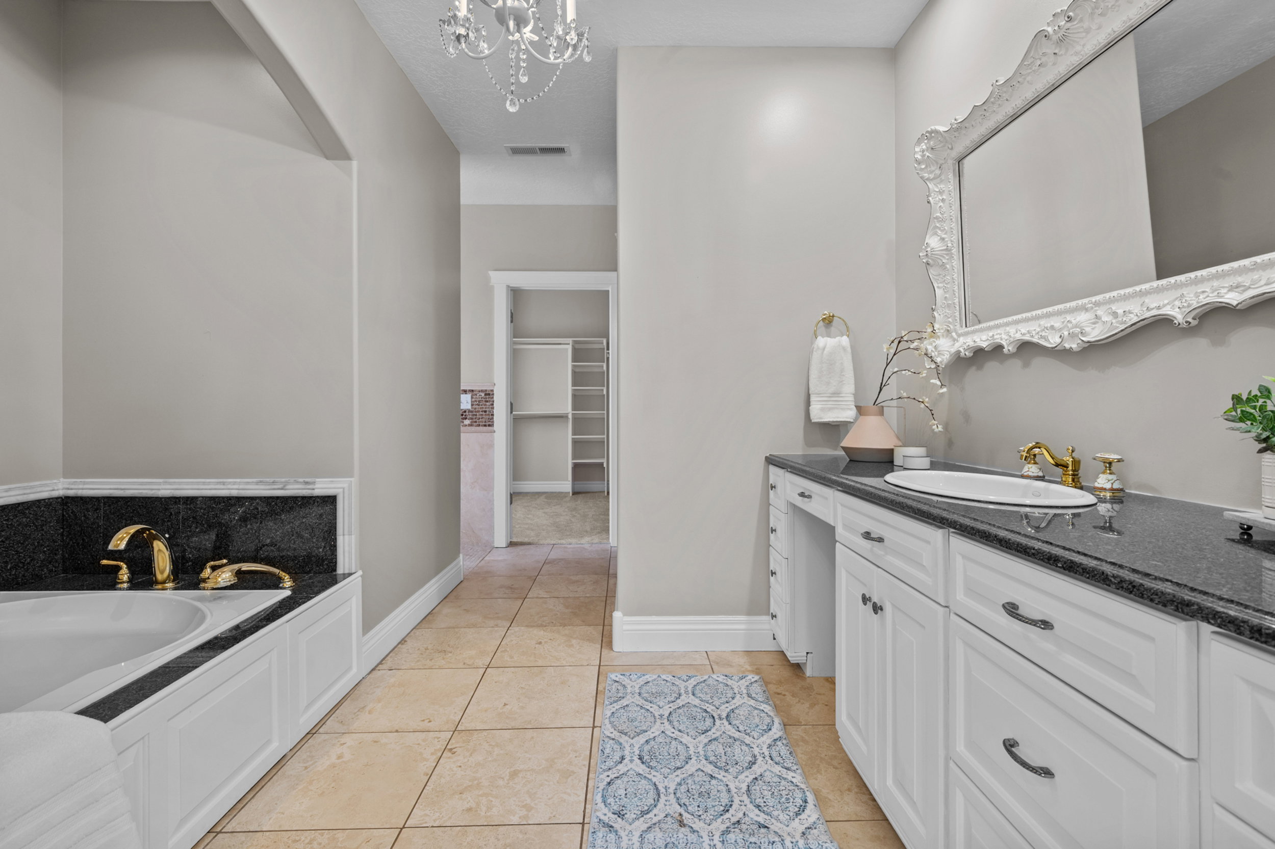 Spacious bathroom featuring a built-in tub with gold-tone fixtures, a dual vanity with a dark countertop, an ornate framed mirror, and tiled flooring