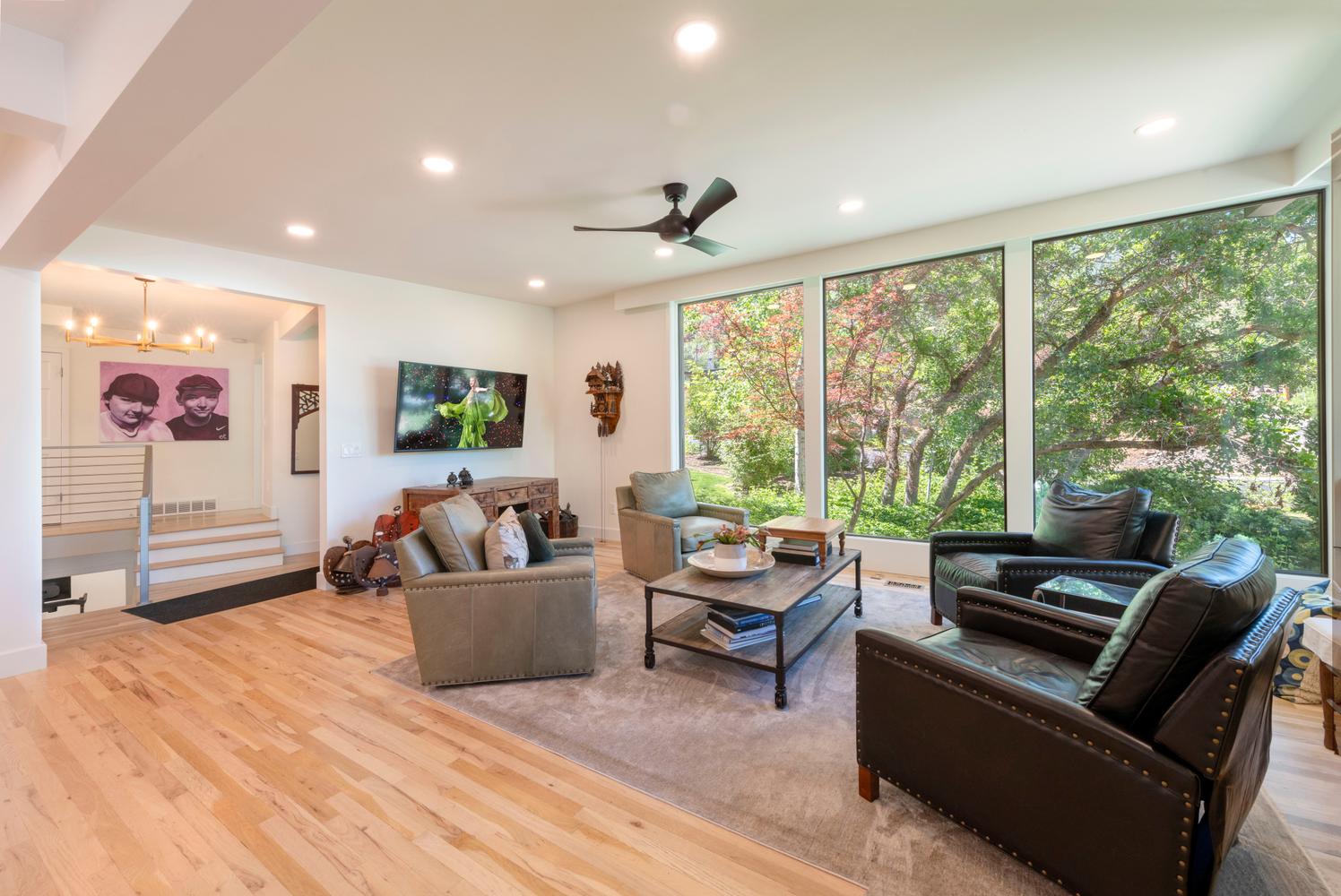 Spacious living area featuring expansive floor-to-ceiling windows, wood-finish flooring, recessed lighting, a contemporary ceiling fan, and a built-in staircase with a metal and glass railing