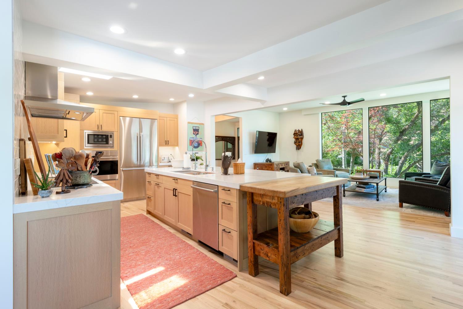 Open-concept kitchen featuring light wood cabinetry, stainless steel appliances, a white countertop island with an undermount sink, and wood-finish flooring extending into the living area with expansive windows