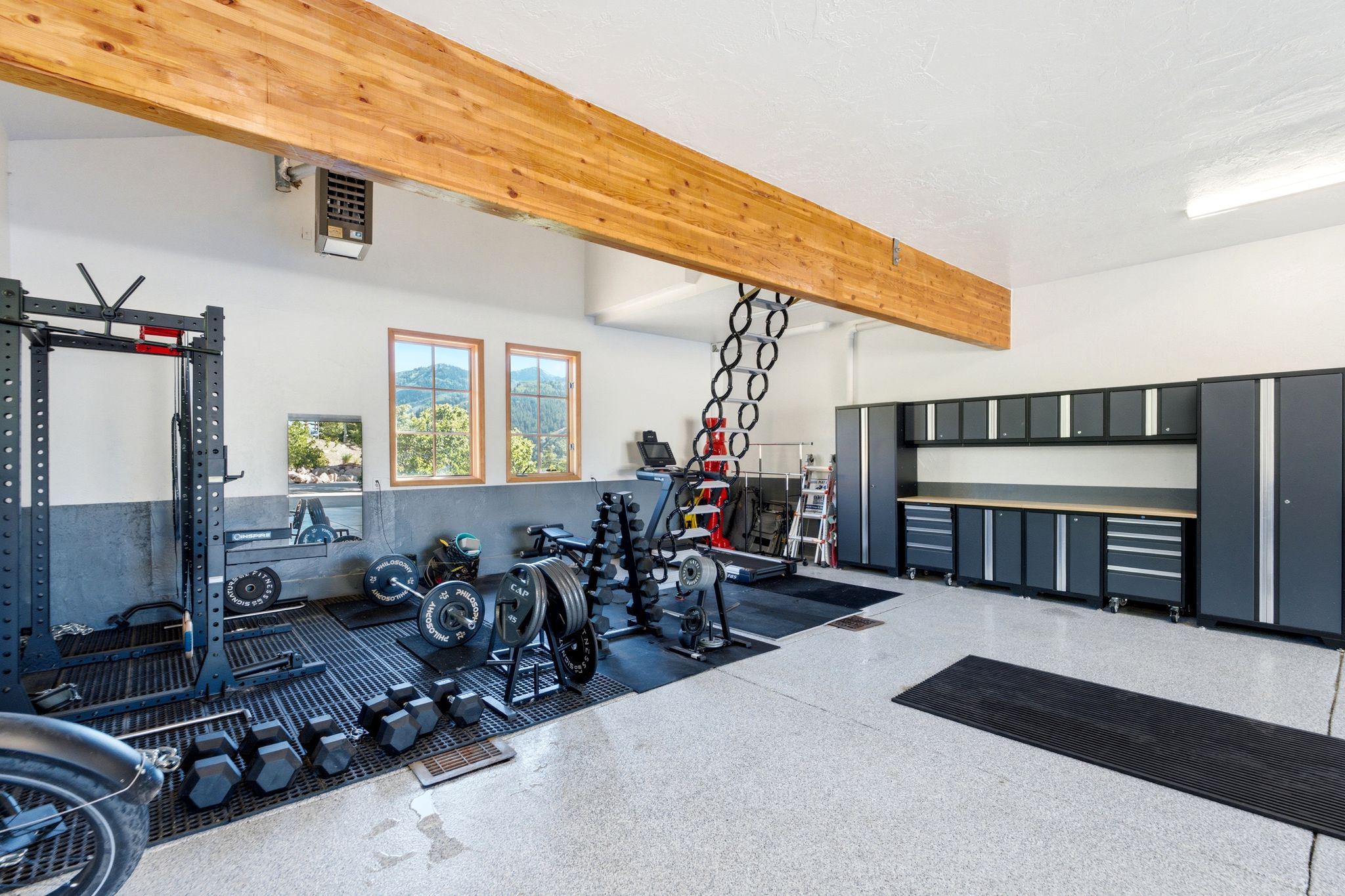 Spacious garage featuring a light wood ceiling beam, light gray speckled epoxy flooring, built-in gray cabinetry with a wood-finish countertop, and multiple windows