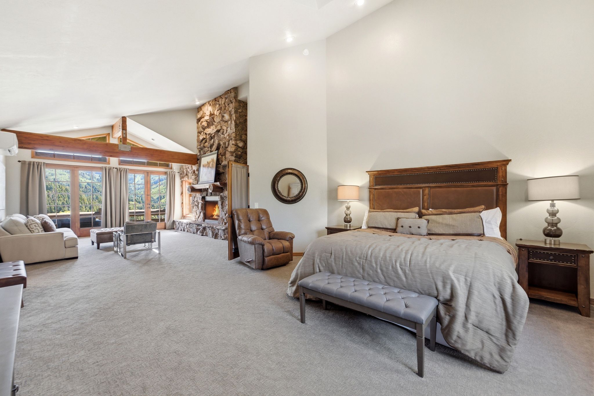 Spacious room featuring vaulted ceilings with exposed wood beams, a stone fireplace, light-colored carpeting, a large bed with a dark wood headboard, and multiple French doors opening to an outdoor area