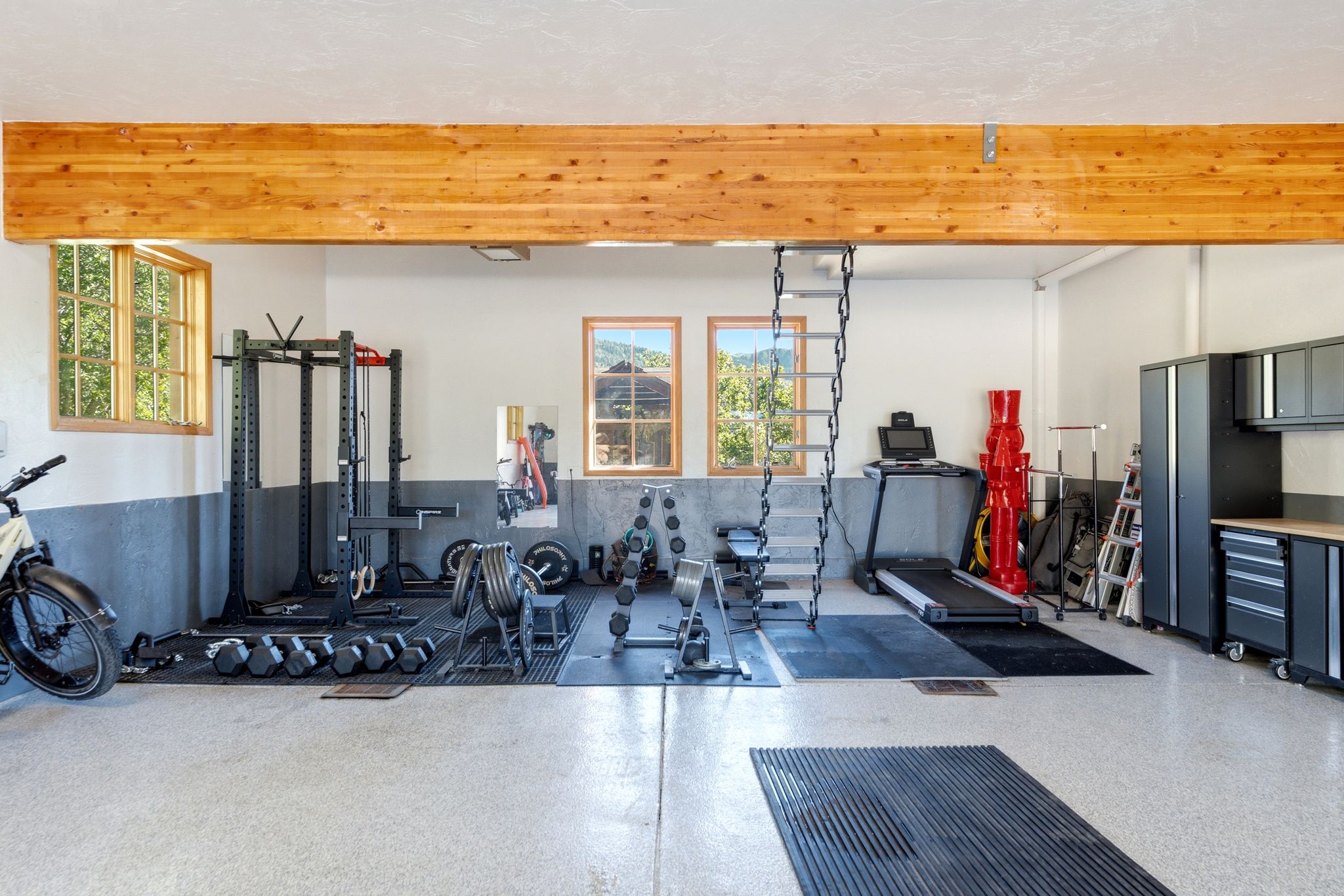 Spacious multi-purpose room featuring an exposed wood beam, three large windows with wood-finish trim, epoxy flooring, and ample built-in storage cabinetry