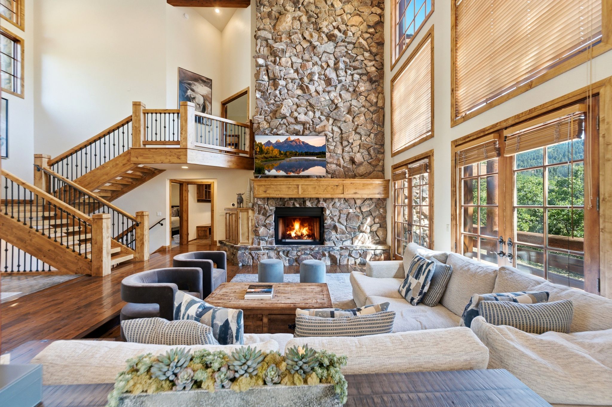 Spacious room featuring a towering stone fireplace, high ceilings, and a grand wooden staircase with wrought iron balusters