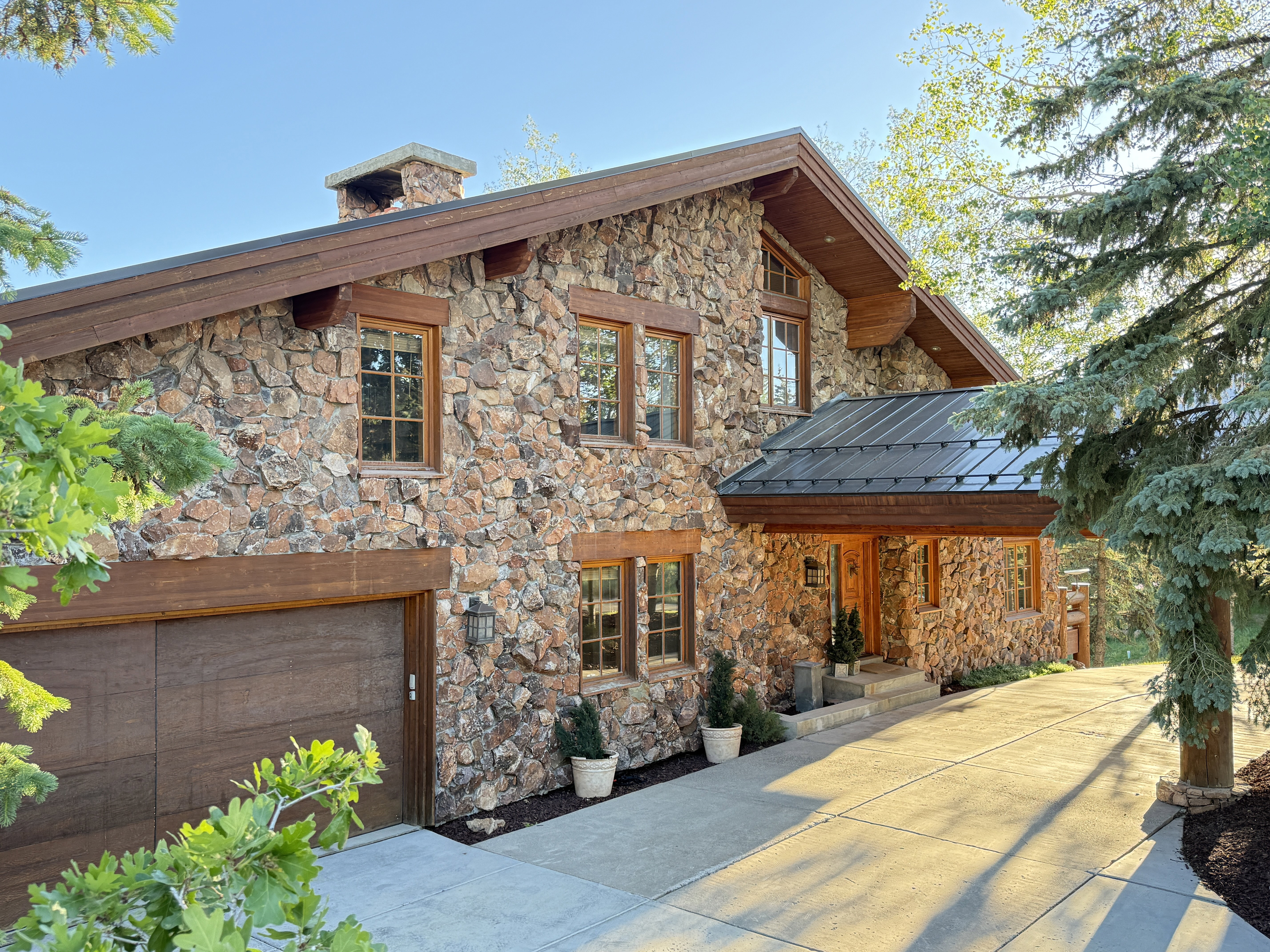 Stone-clad exterior featuring a wood-finish garage door, prominent chimney, dark metal roof over the entry, and a concrete driveway