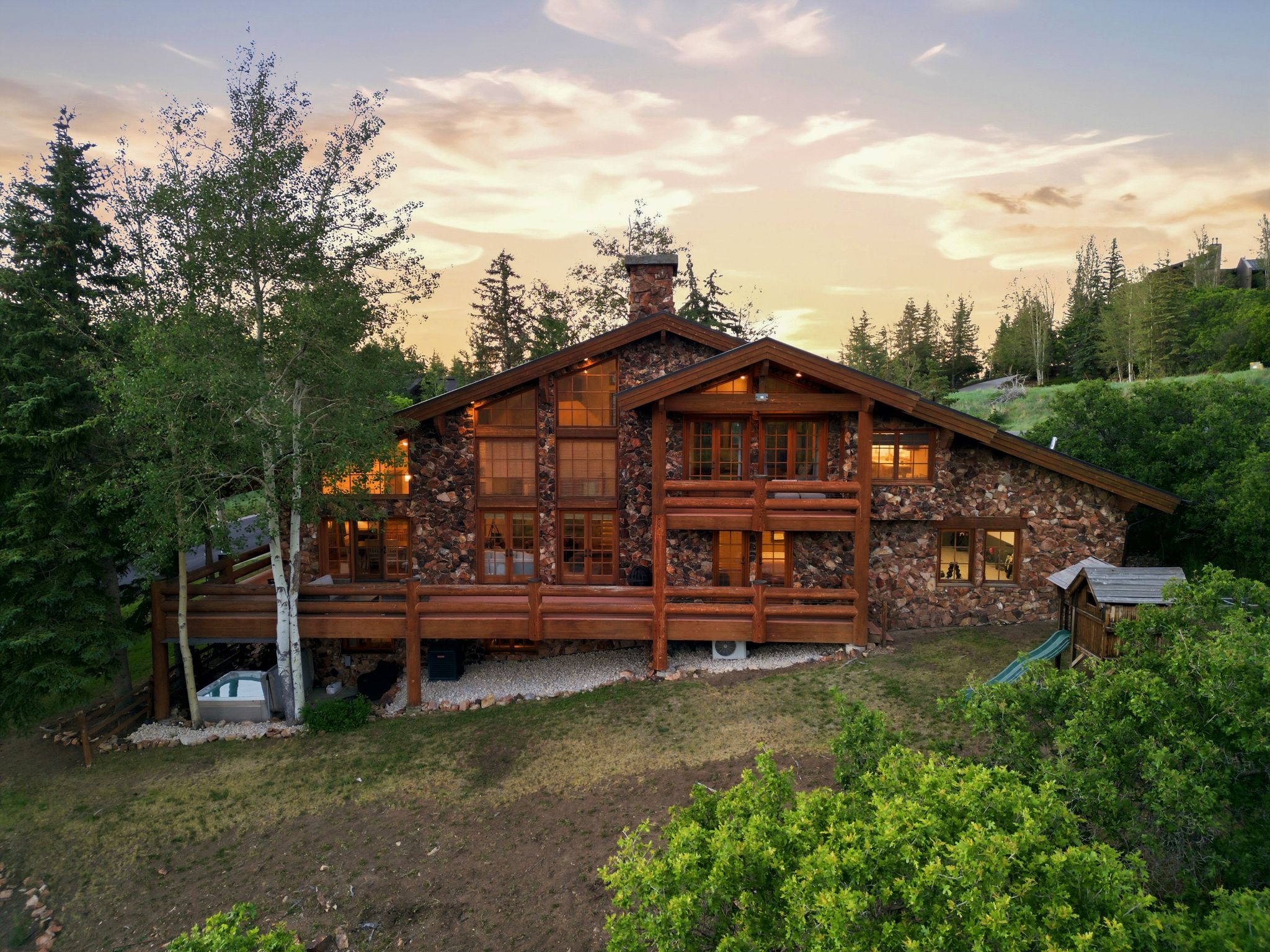 Stone and wood-sided exterior featuring multiple balconies, a large deck, a built-in hot tub, and mature trees
