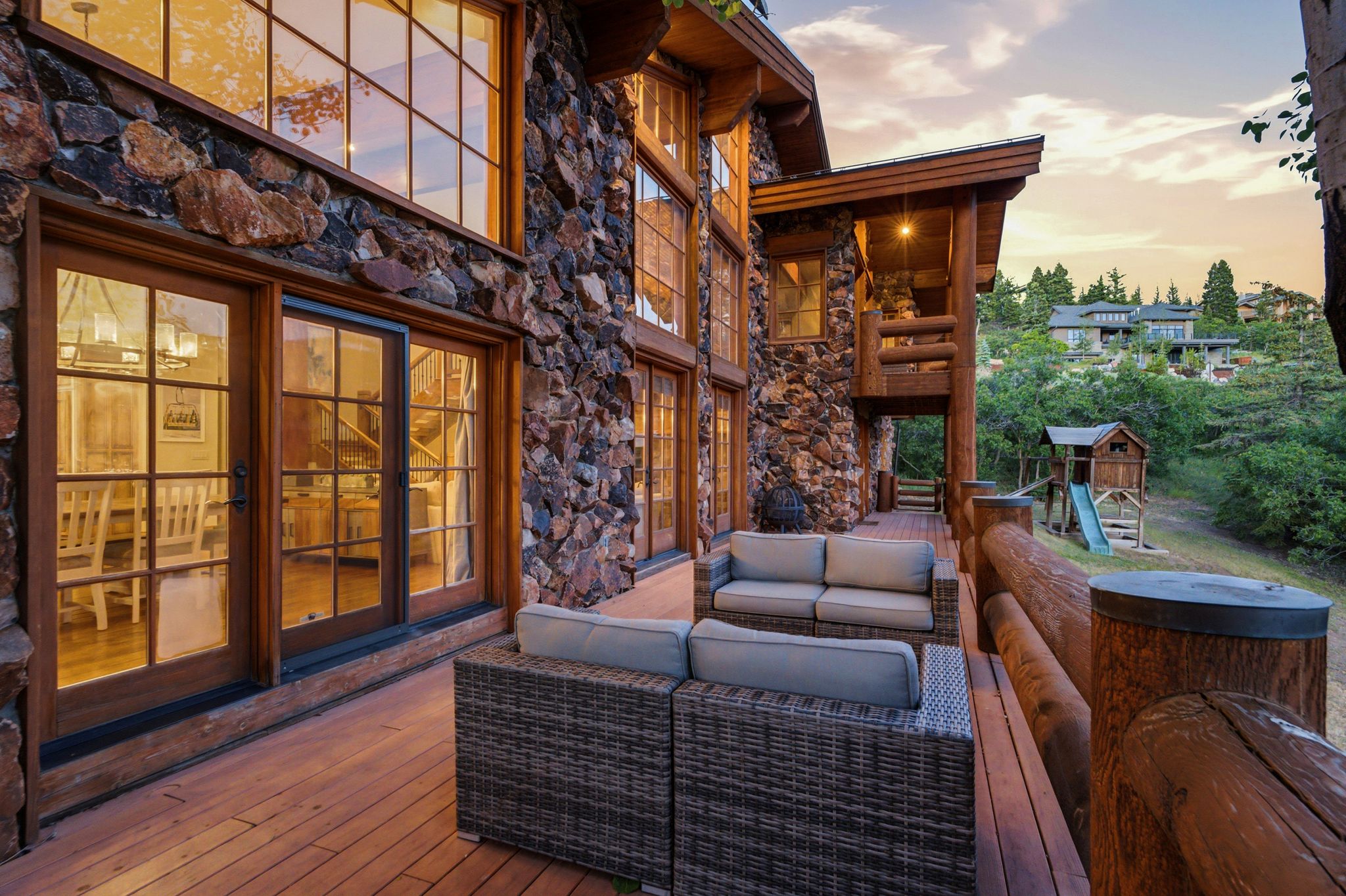 Spacious outdoor wood-finish deck with log railing, featuring a stone exterior, large wood-framed windows, and French doors
