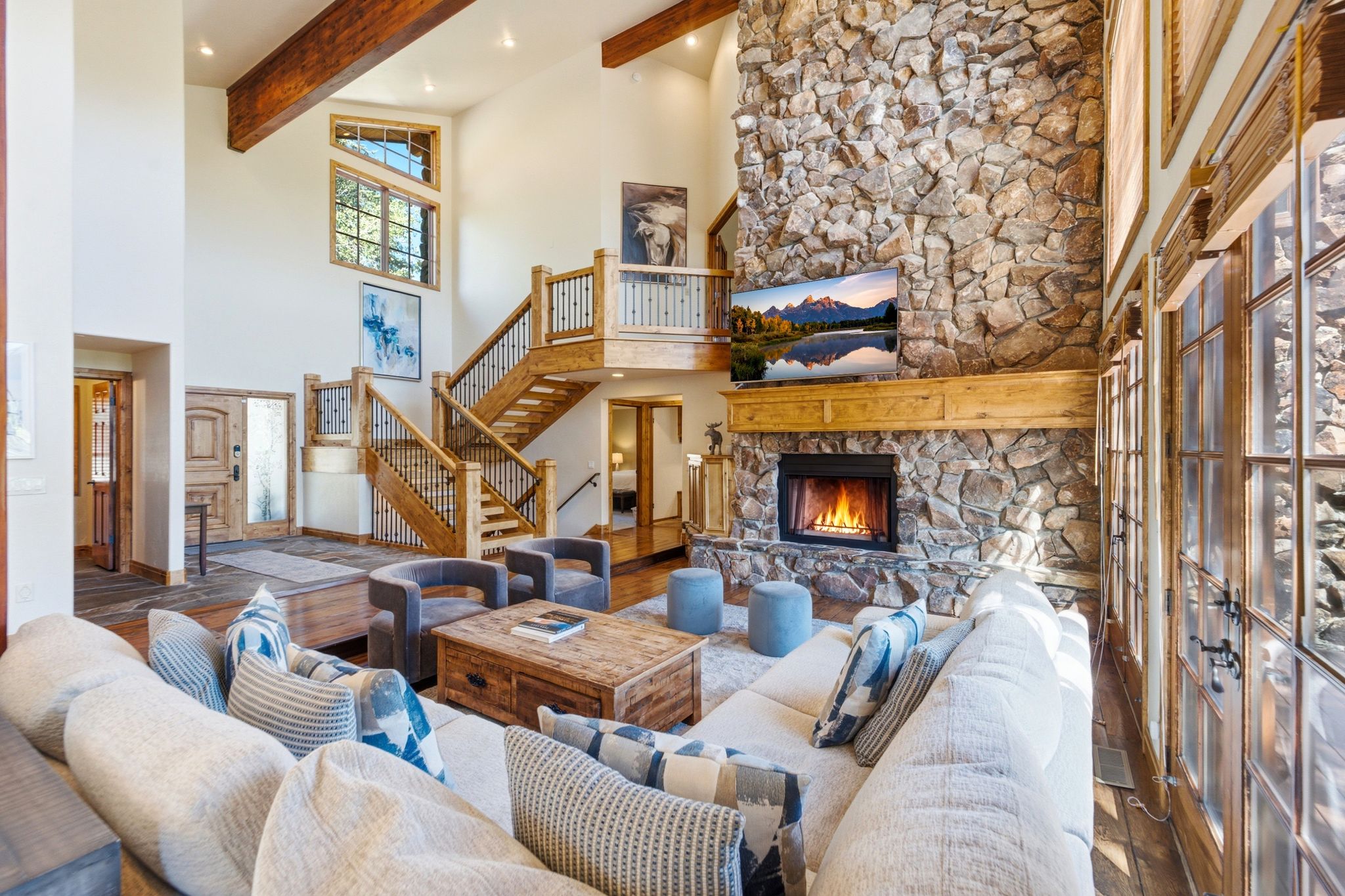 Spacious living area featuring a stone fireplace with a wood mantle, high ceilings with exposed wood beams, and large multi-pane windows