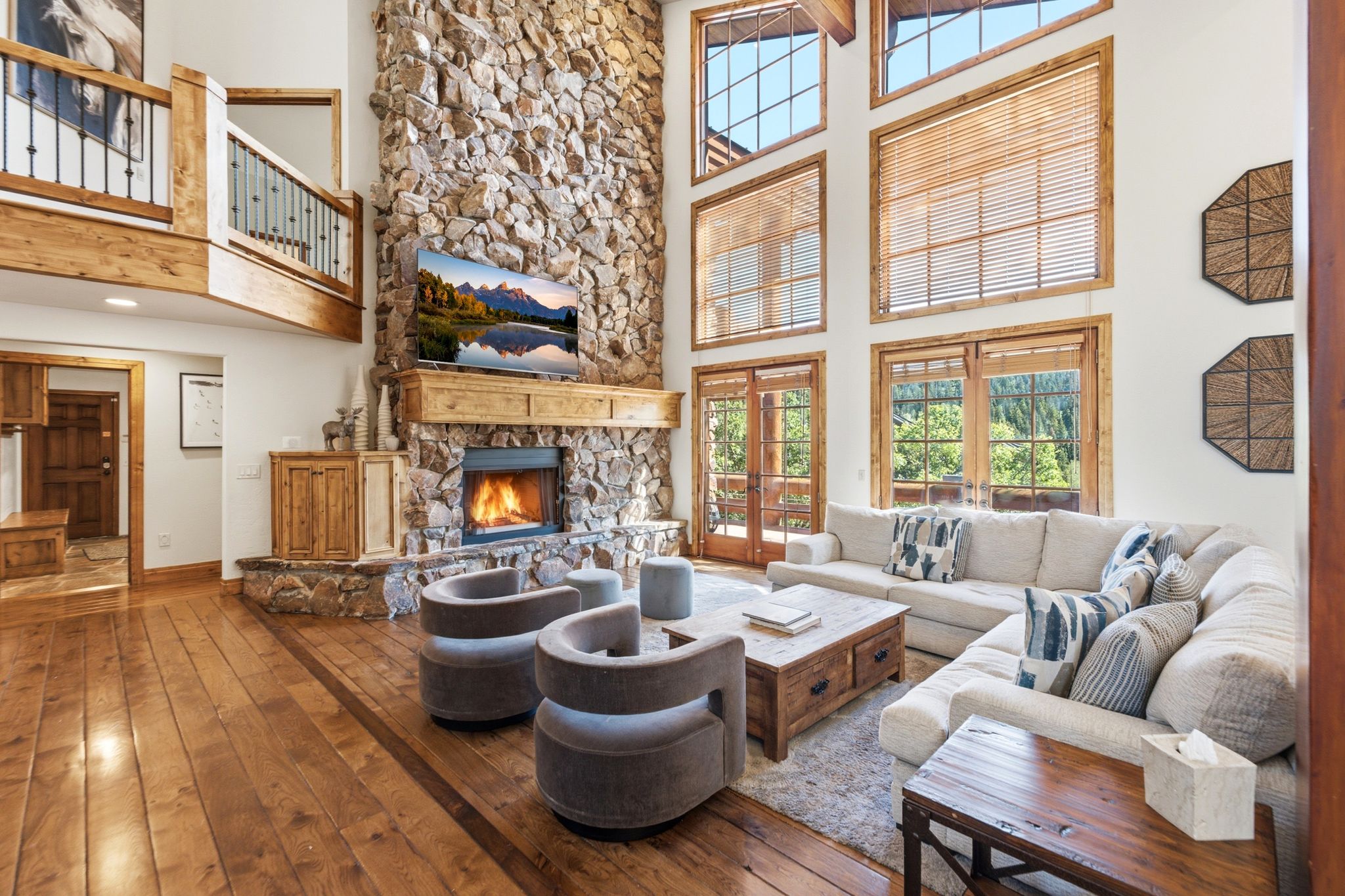 Great room featuring a towering stone fireplace, high ceilings, large multi-pane windows, wood-finish flooring, and a second-story balcony with iron railings