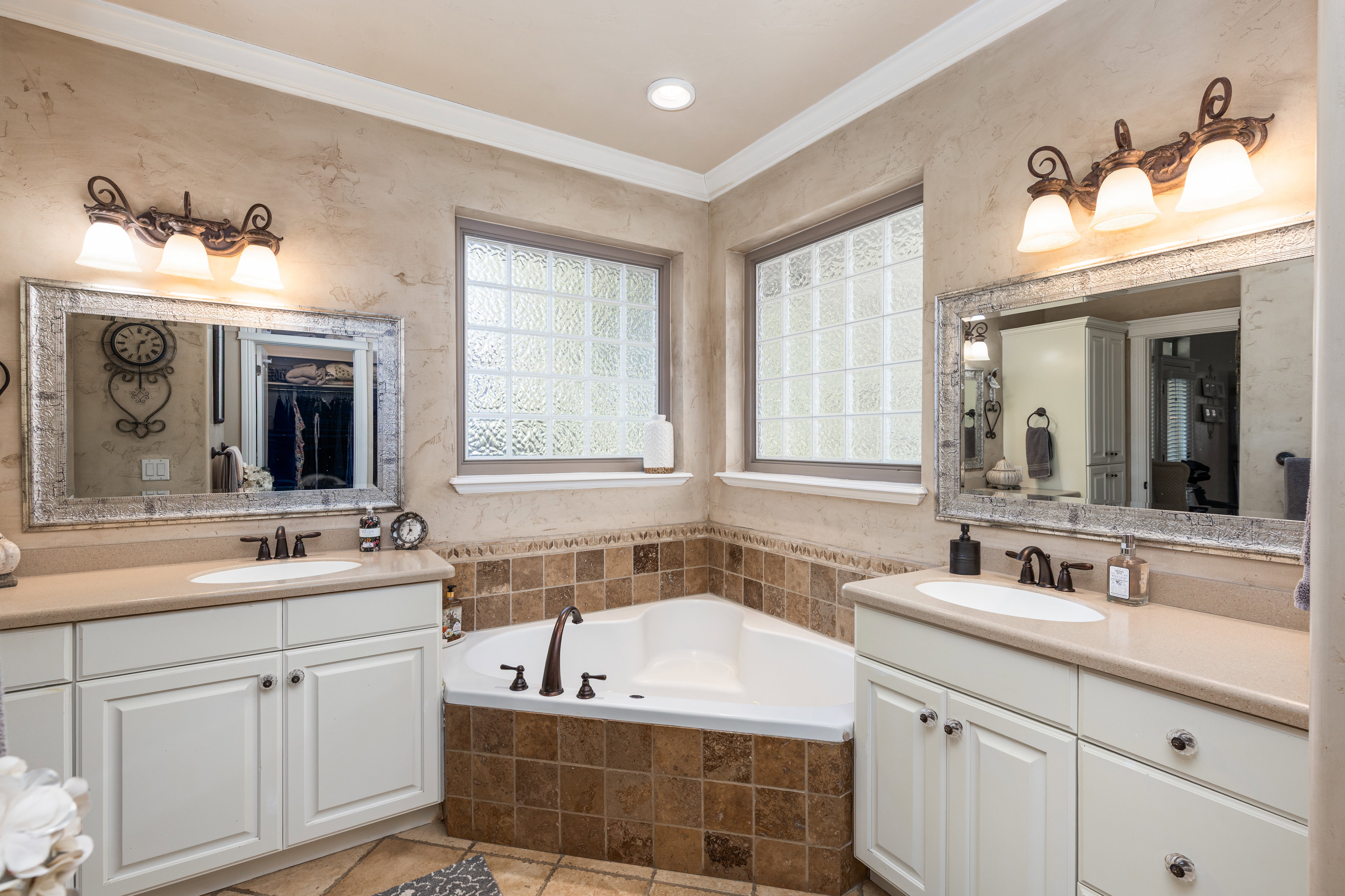 Dual vanity sinks with cream-colored countertops, an integrated corner soaking tub, textured wall finish, and two frosted glass block windows