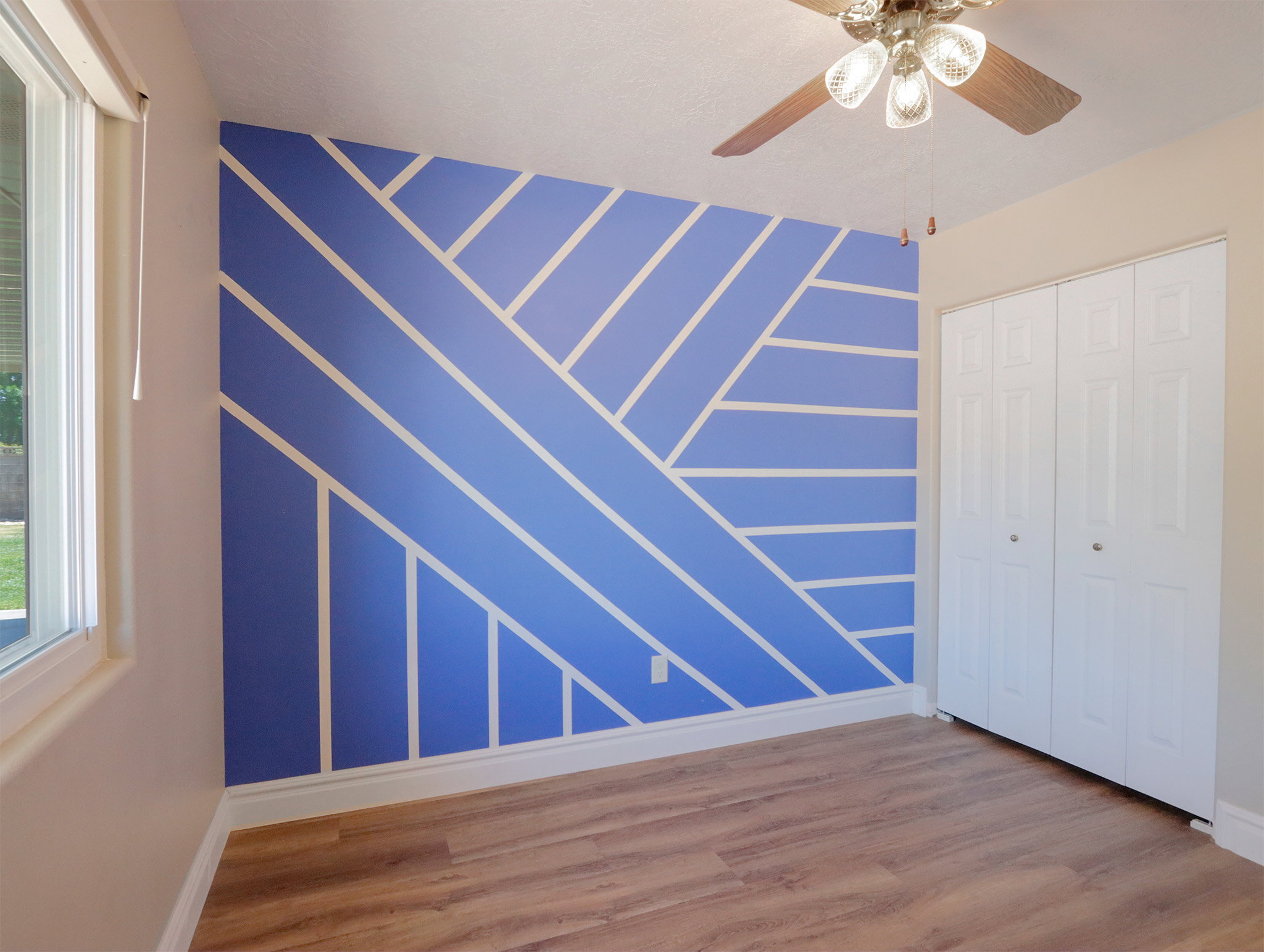 Feature wall with geometric pattern, wood-finish flooring, ceiling fan with light fixture, white bi-fold closet doors, and a window providing natural light