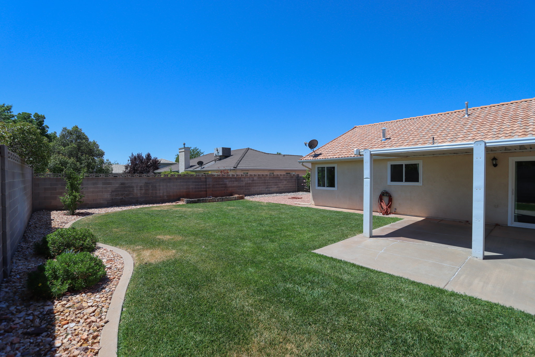 Expansive grass backyard featuring a concrete patio, perimeter block wall fencing, and a decorative rock border with shrubbery