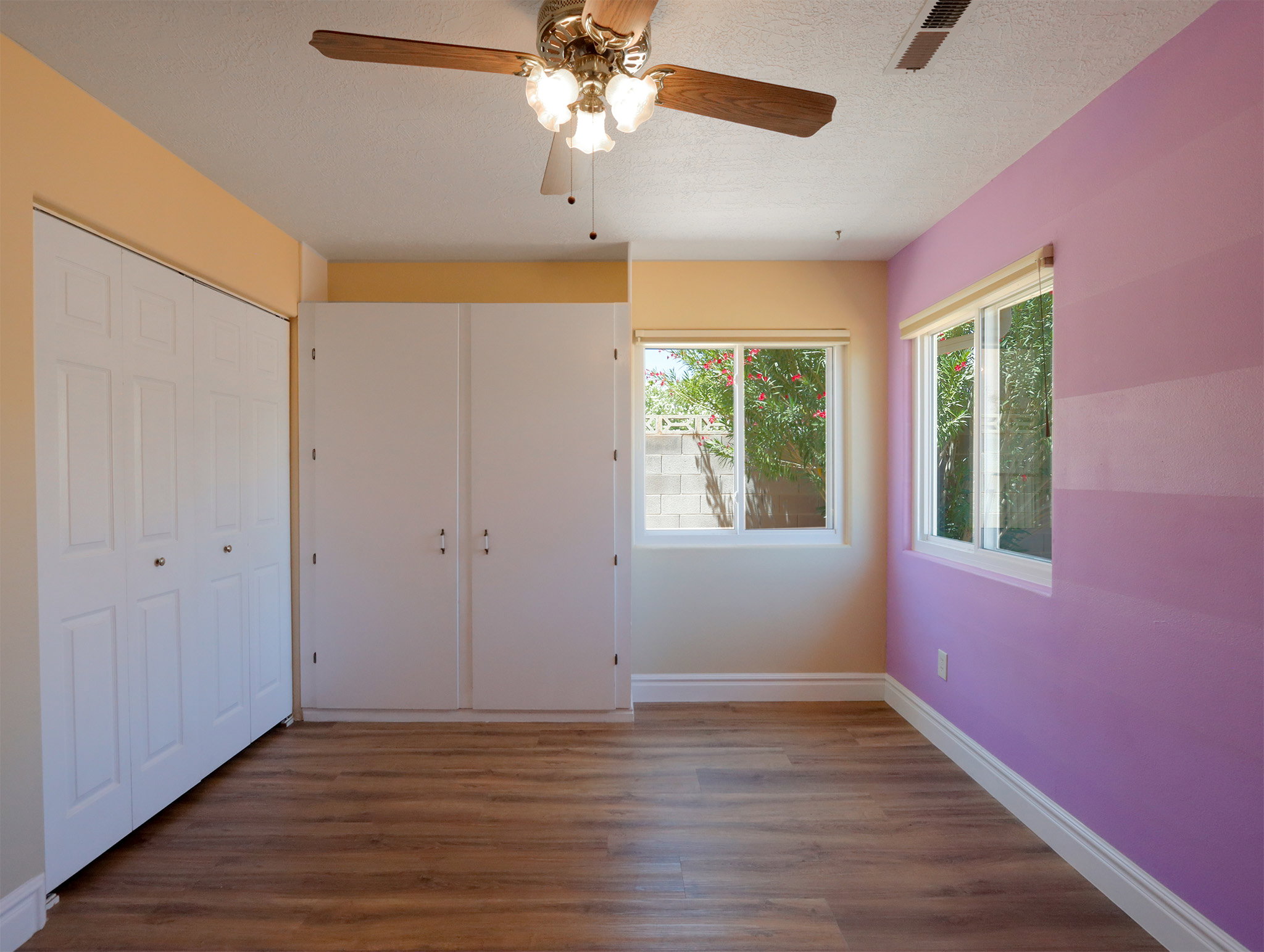 Room featuring wood-finish flooring, two large windows, a ceiling fan with integrated lighting, and multiple storage closets with panel doors