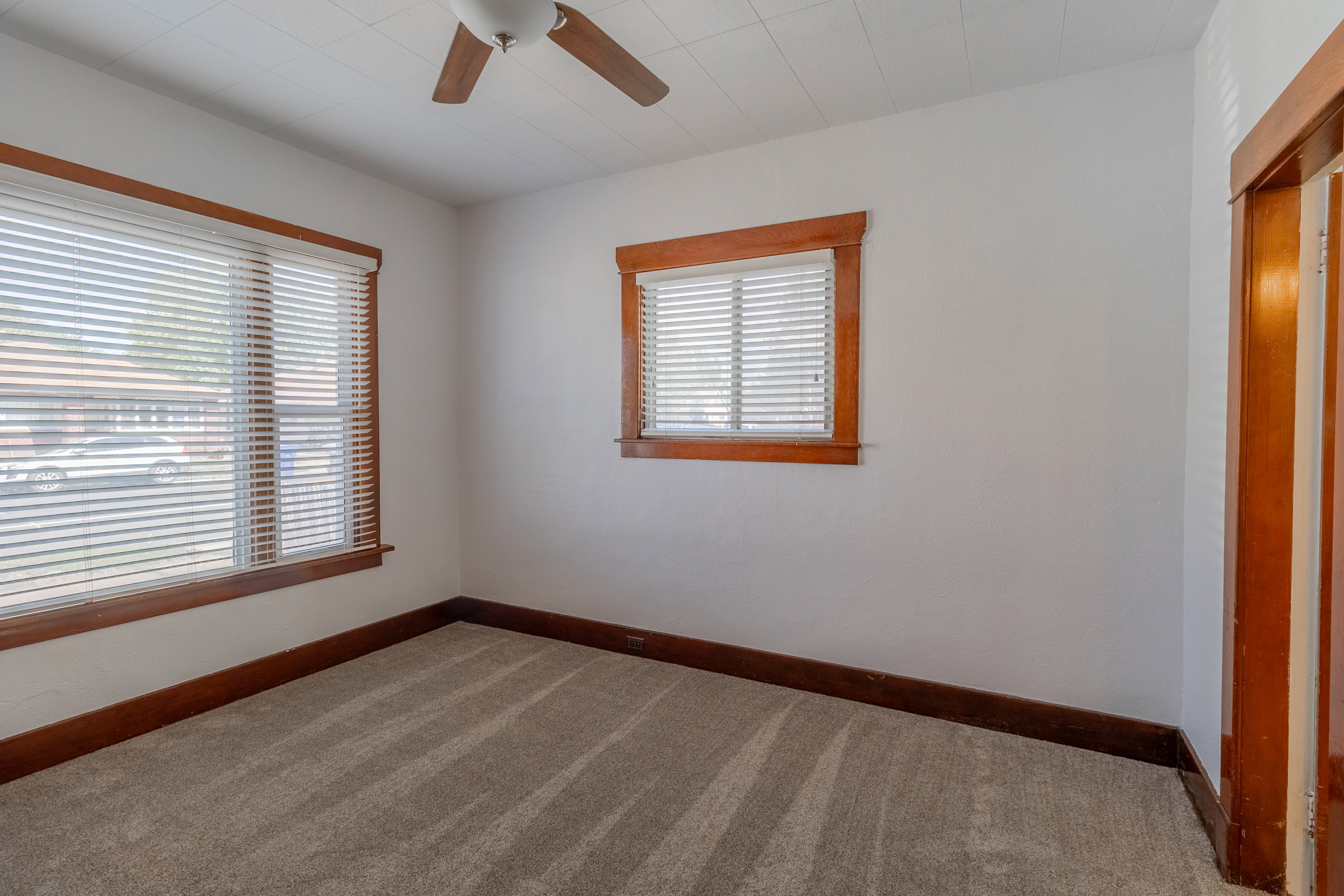 Carpeted room featuring white walls, two windows with blinds, a ceiling fan, and wood trim around windows, baseboards, and doorway
