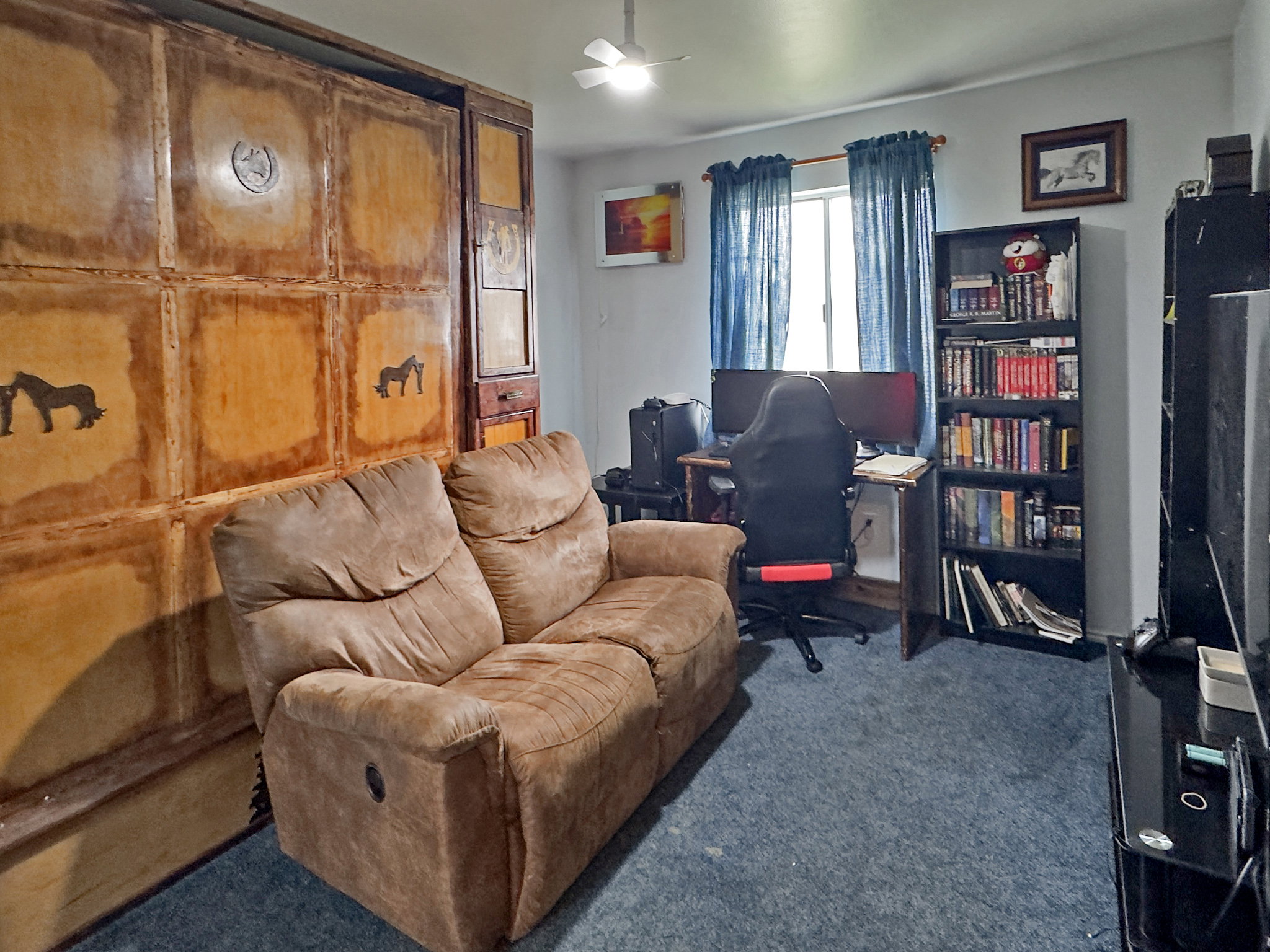 Room featuring a unique built-in wooden wall unit with decorative accents, blue carpeting, and a ceiling fan
