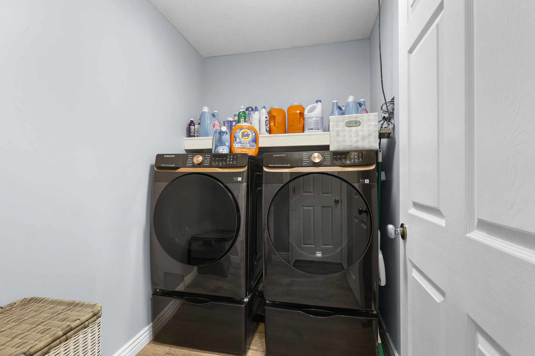 Dedicated laundry area featuring a front-loading washer and dryer set on pedestals, overhead shelving, light blue wall paint, white trim, and wood-finish flooring