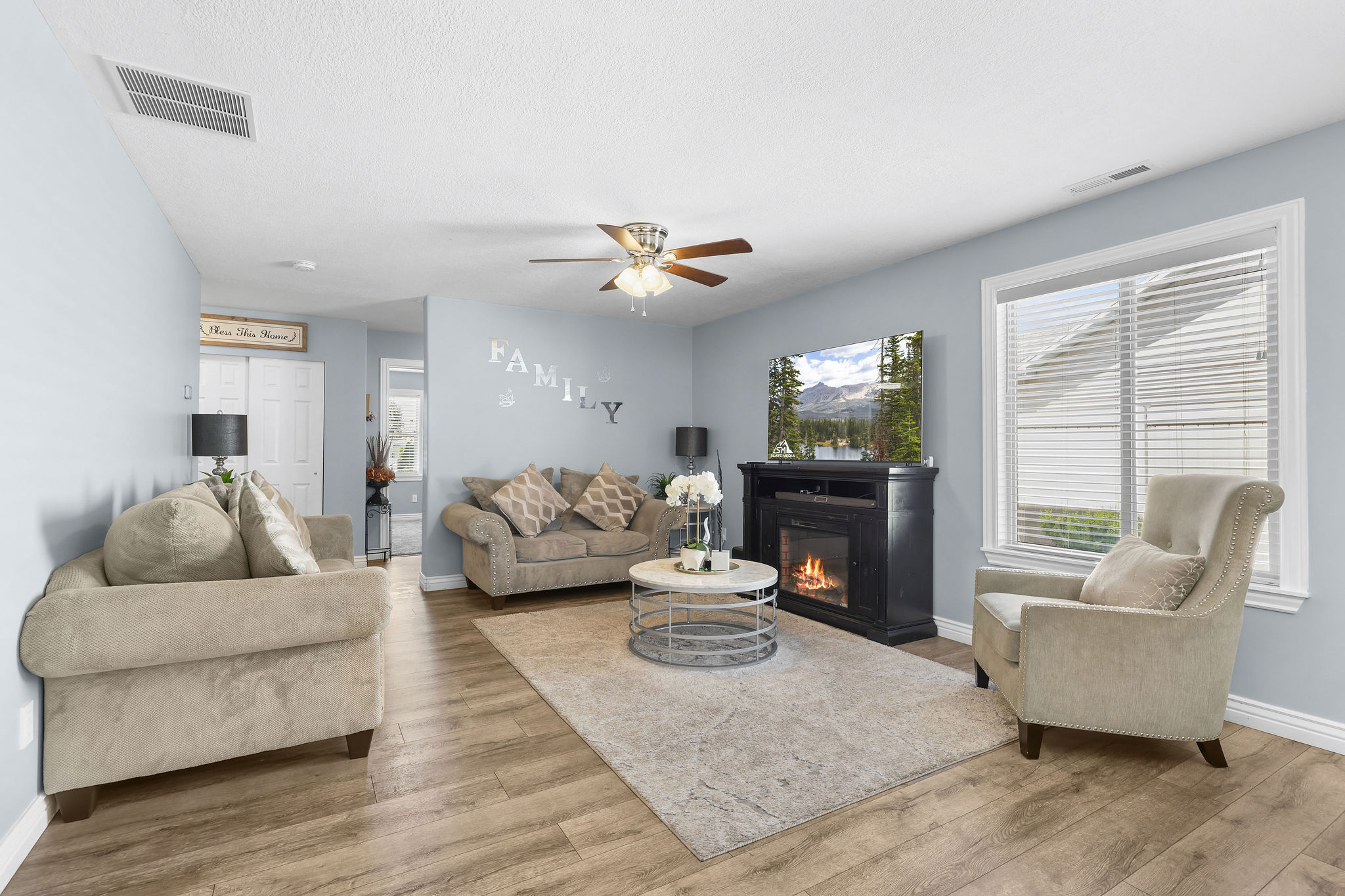 Living area featuring wood-finish flooring, a ceiling fan, and a large window with blinds