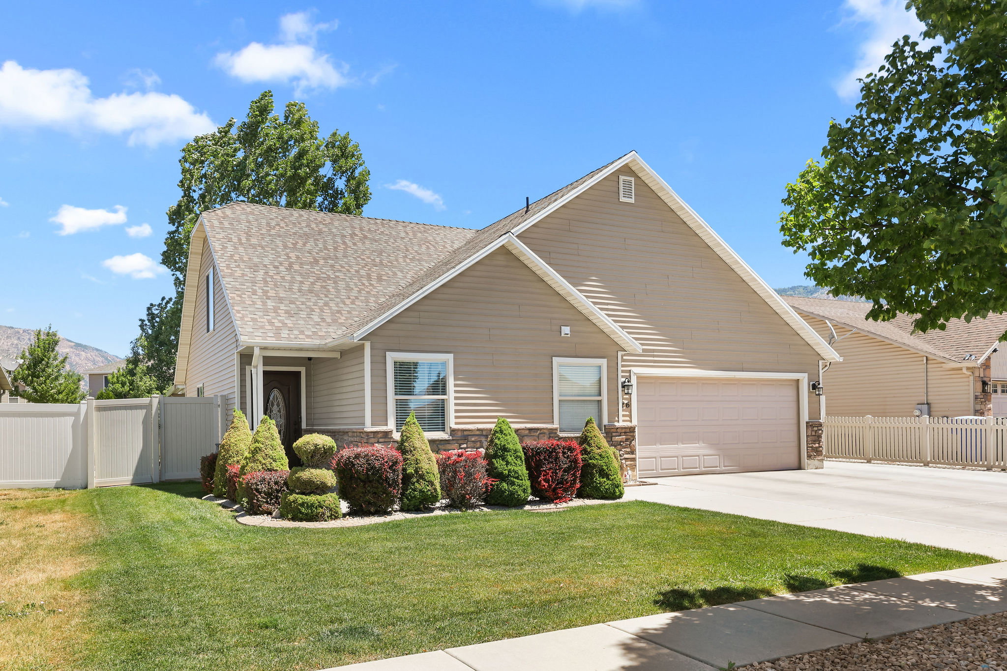 Single-story residence featuring light brown horizontal siding with stone veneer accents, a prominent front gable, an attached two-car garage, and a manicured front lawn with varied shrubbery