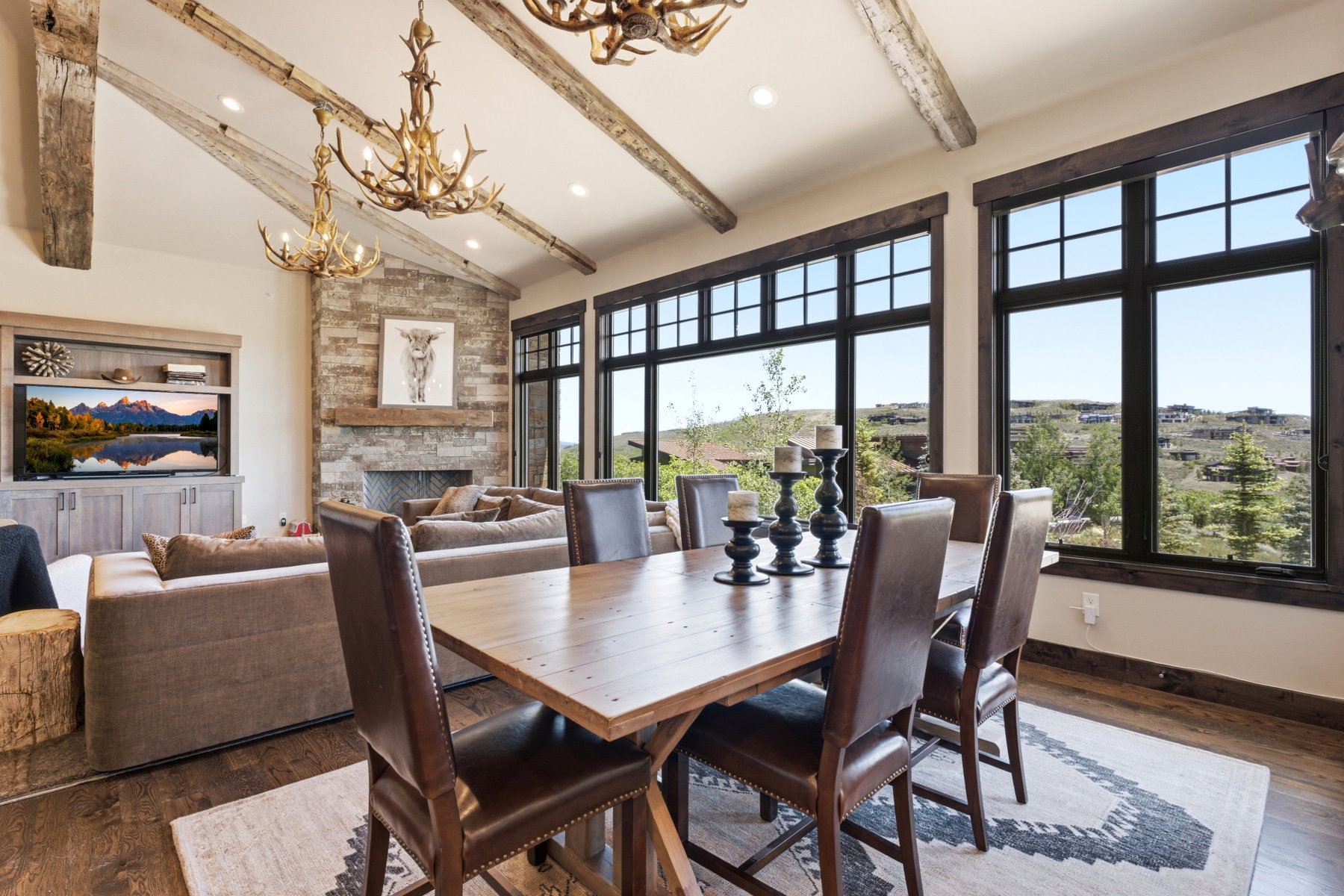 Open-concept living and dining area featuring a vaulted ceiling with exposed wood beams, a stone-faced fireplace with a wood mantel, a built-in media center, and expansive windows with dark frames
