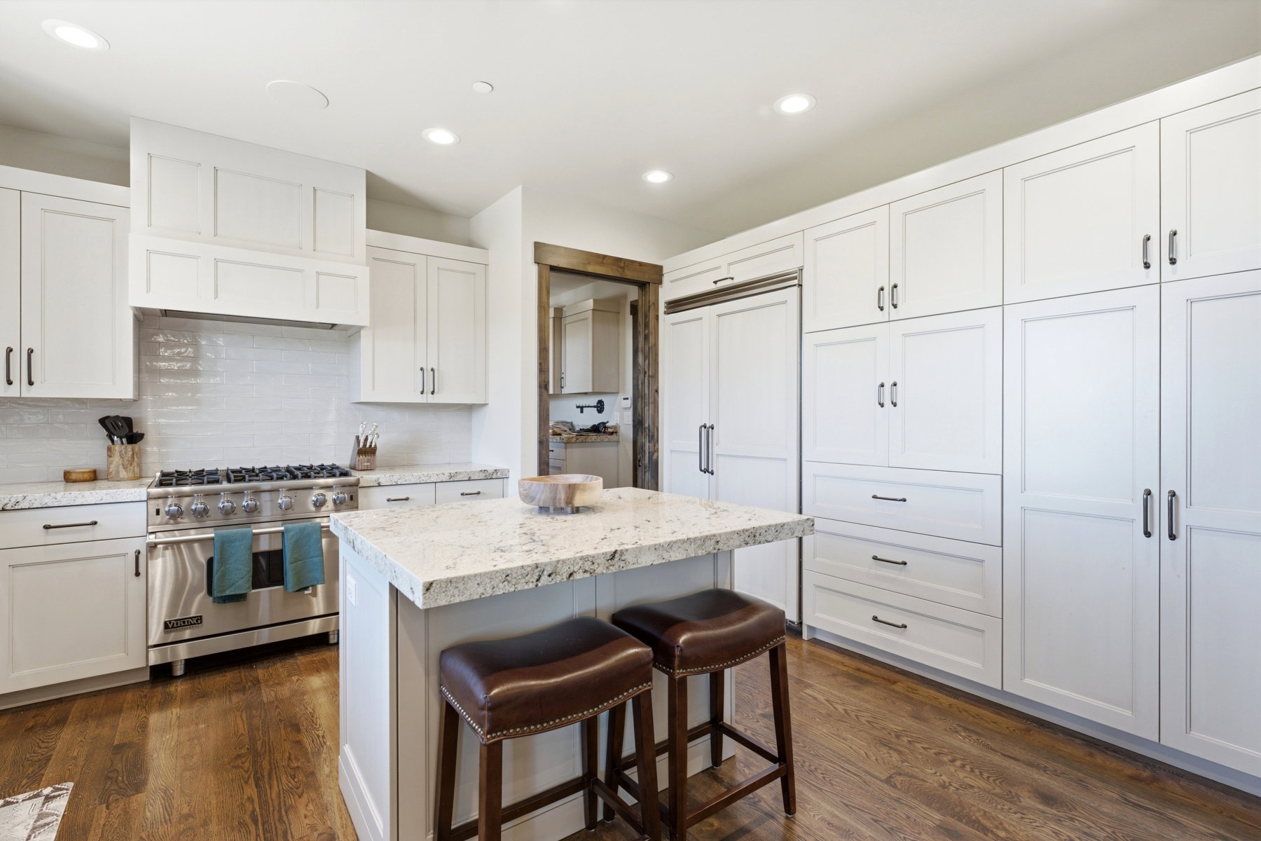 Gourmet kitchen featuring a built-in stainless steel range, white shaker cabinetry, an integrated panel-ready refrigerator, wood-finish flooring, and a central island with a stone countertop