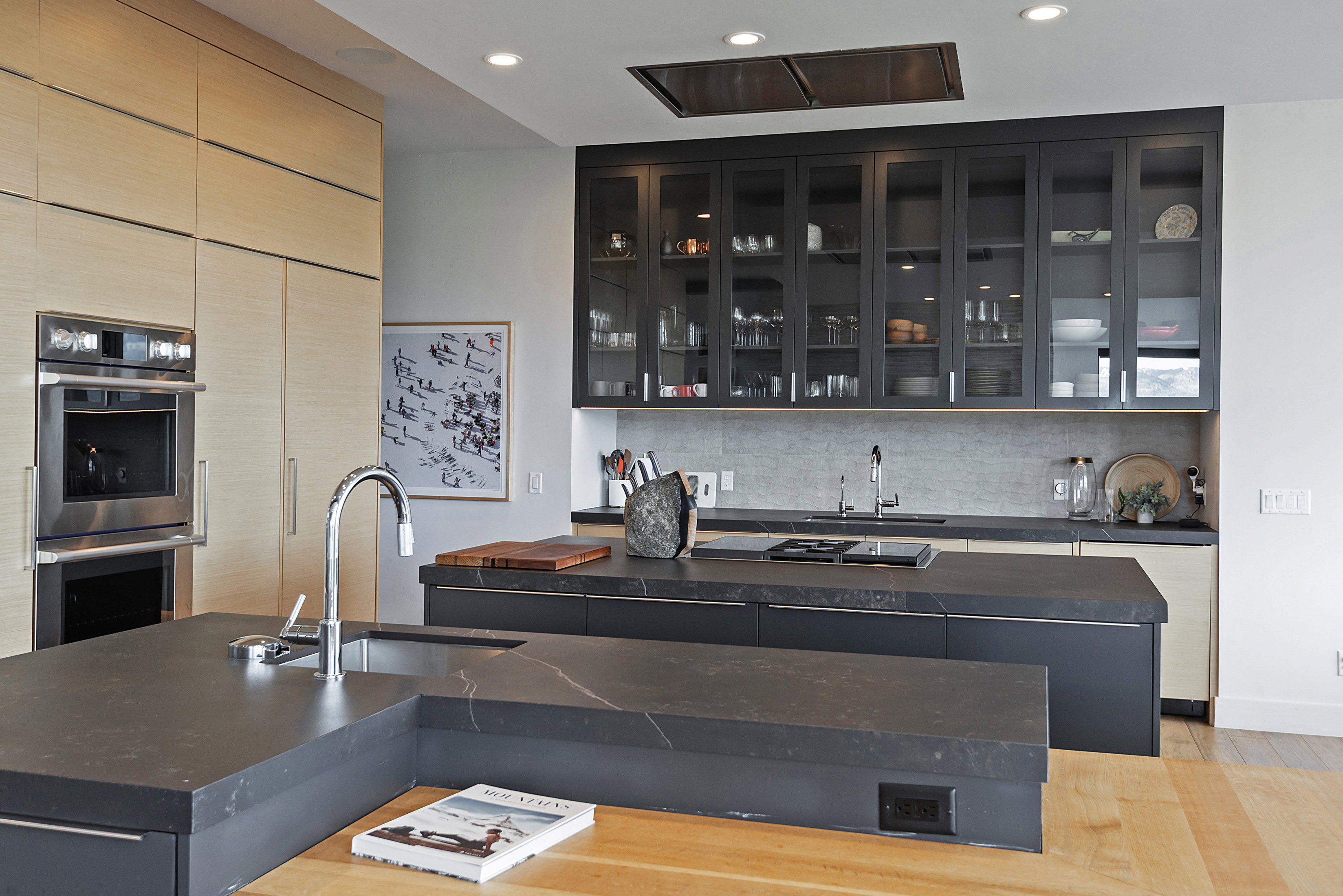 Contemporary kitchen featuring two islands with dark countertops, sleek light wood-finish cabinetry, built-in stainless steel oven, a stainless steel sink with a gooseneck faucet, and dark glass-front upper cabinets