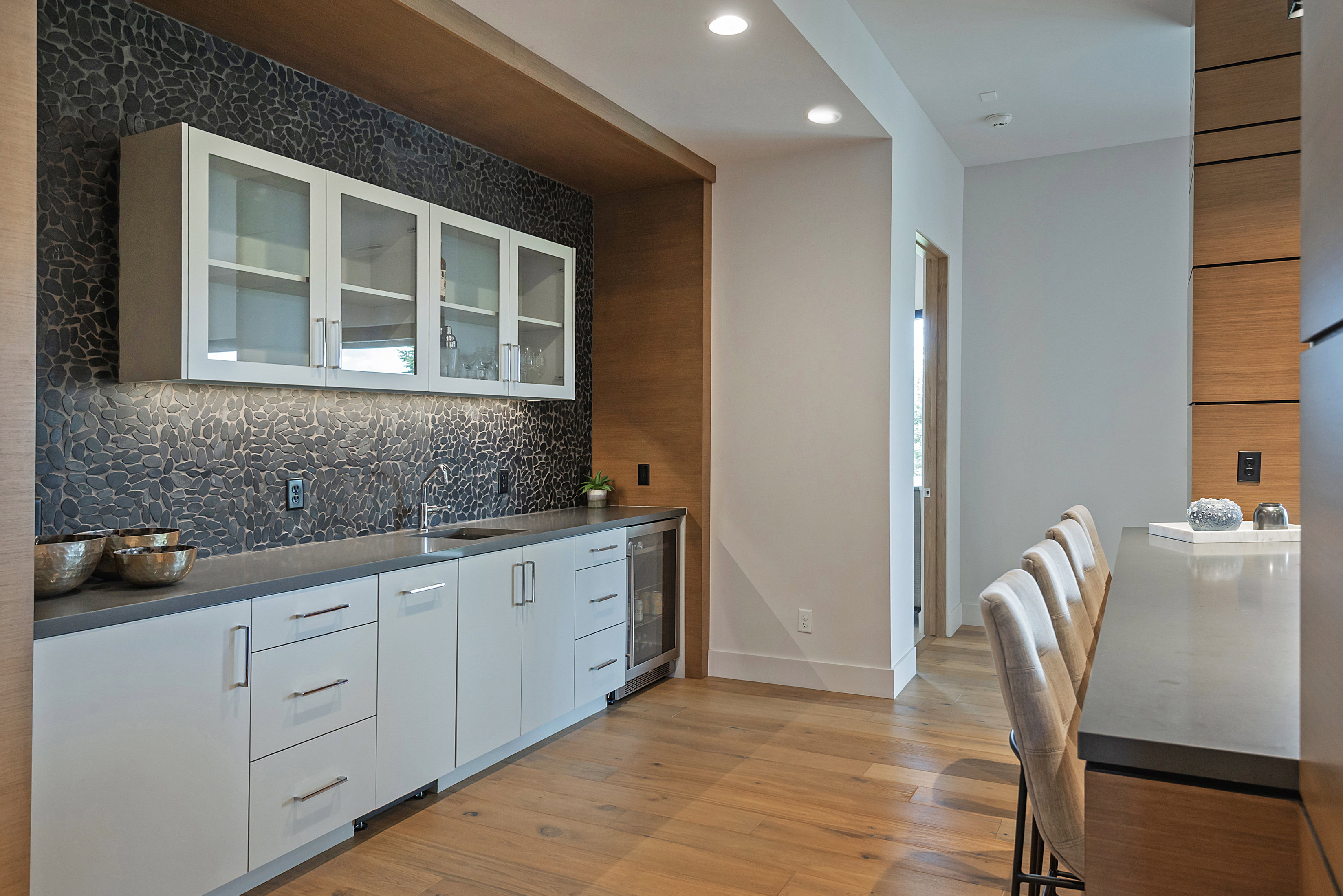 Built-in wet bar featuring a textured dark stone backsplash, under-cabinet lighting, and a stainless steel beverage refrigerator