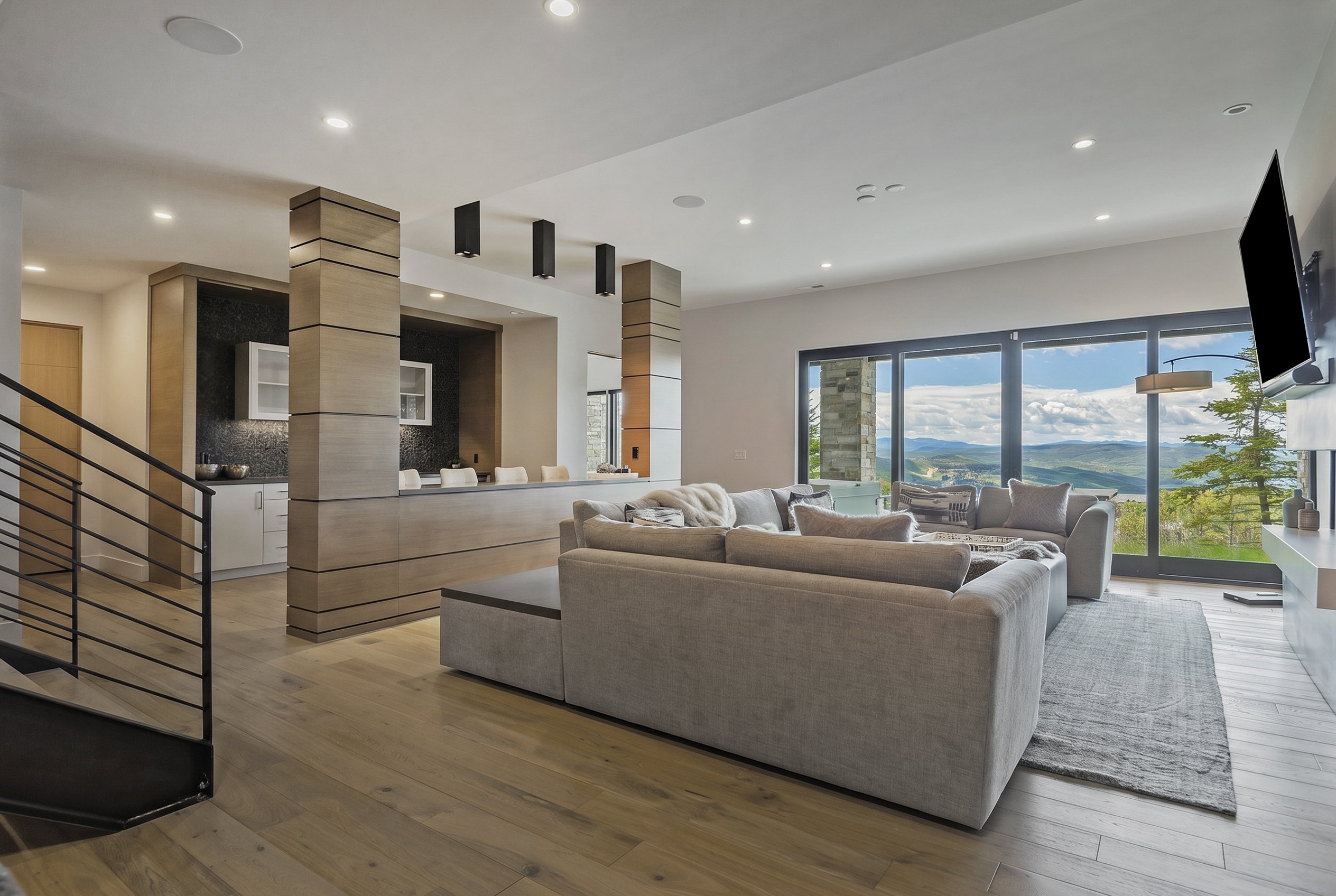 Open-concept living area with wood-finish flooring, a contemporary wet bar featuring dark tile backsplash, and expansive floor-to-ceiling sliding glass doors revealing mountain scenery