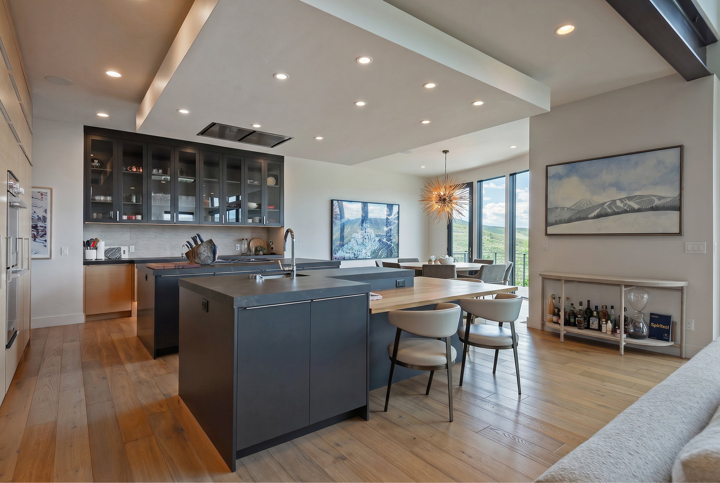 Spacious kitchen area featuring dual islands with integrated seating, contemporary dark cabinetry, light wood-finish flooring, recessed lighting, and large windows capturing an expansive outdoor landscape