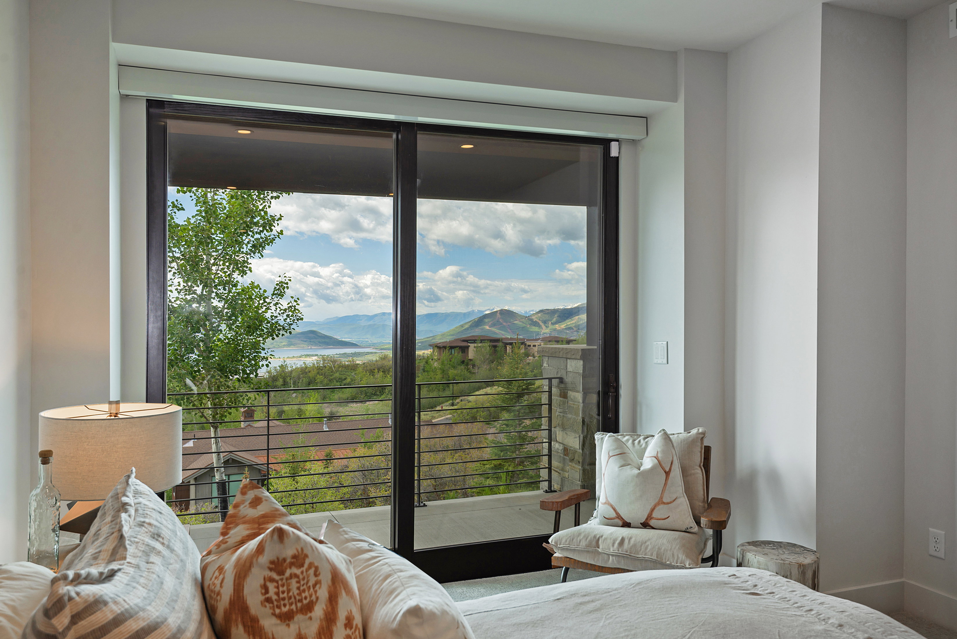 Expansive glass sliding doors open to a balcony with metal railings, offering mountain and lake scenery