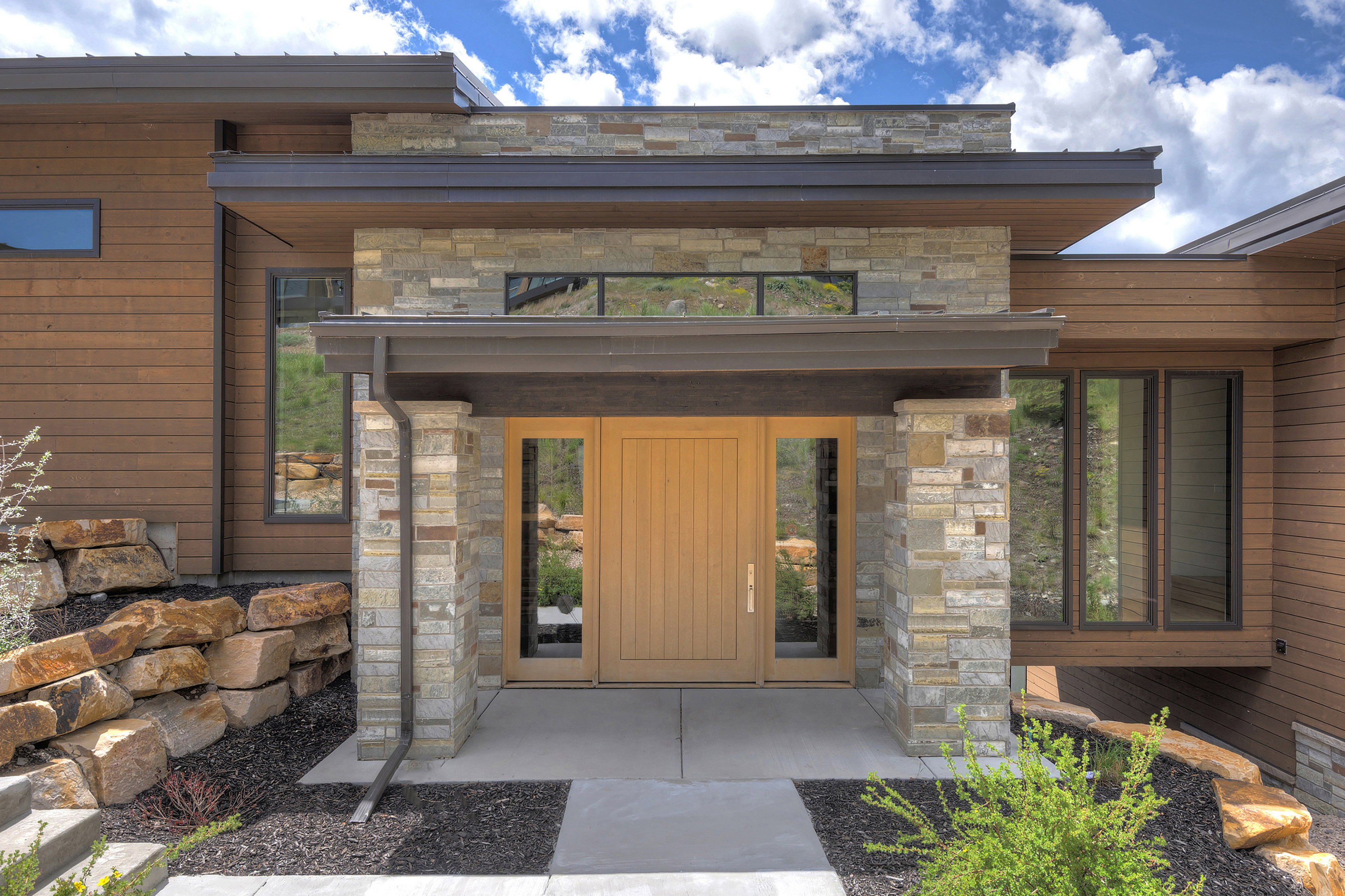 Contemporary exterior featuring a natural wood-finish door, stone accents, horizontal wood siding, and large windows