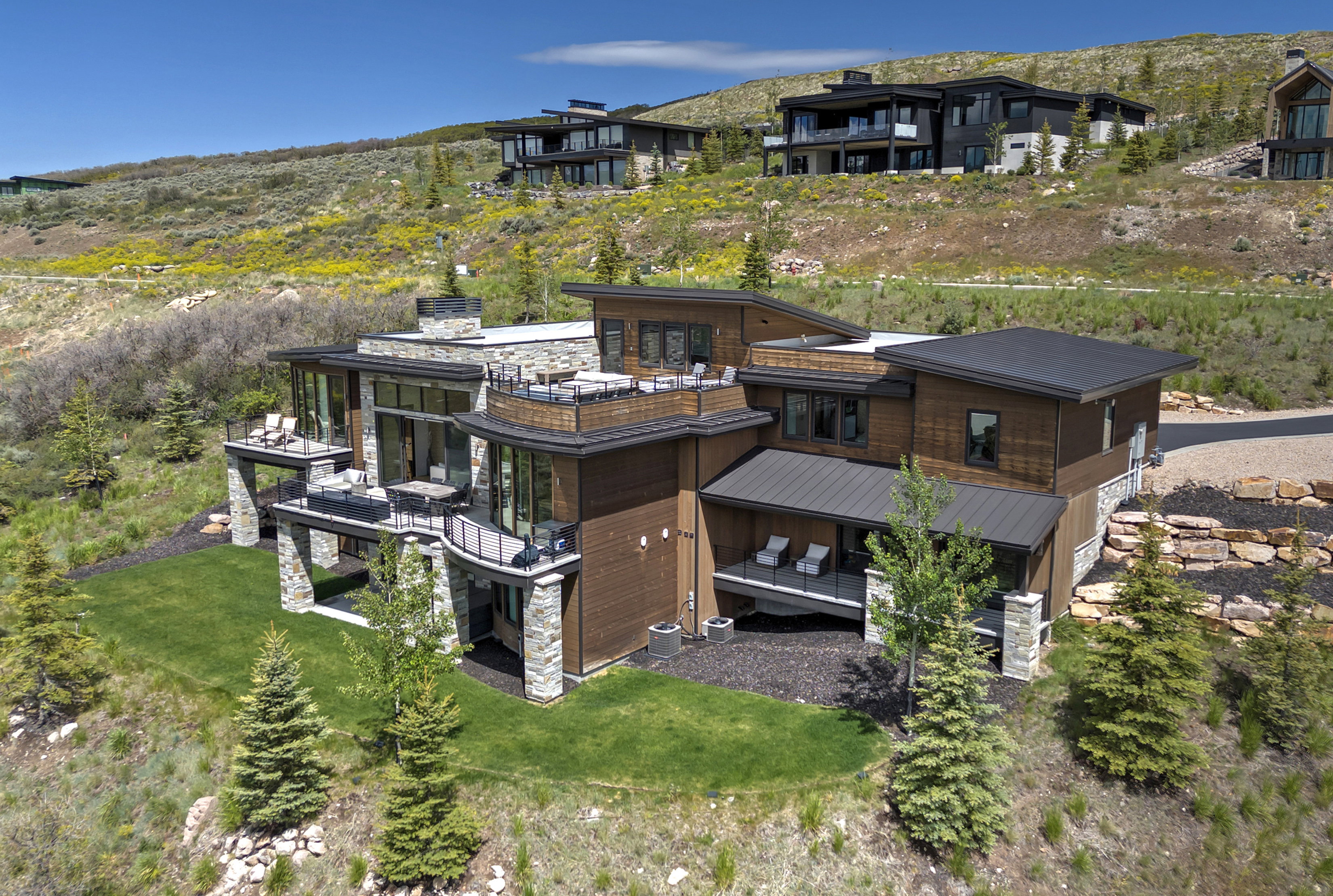 Modern multi-level residence featuring extensive stone and wood siding, multiple exterior balconies with glass railings, a flat roofline, and integrated landscaping with mature trees