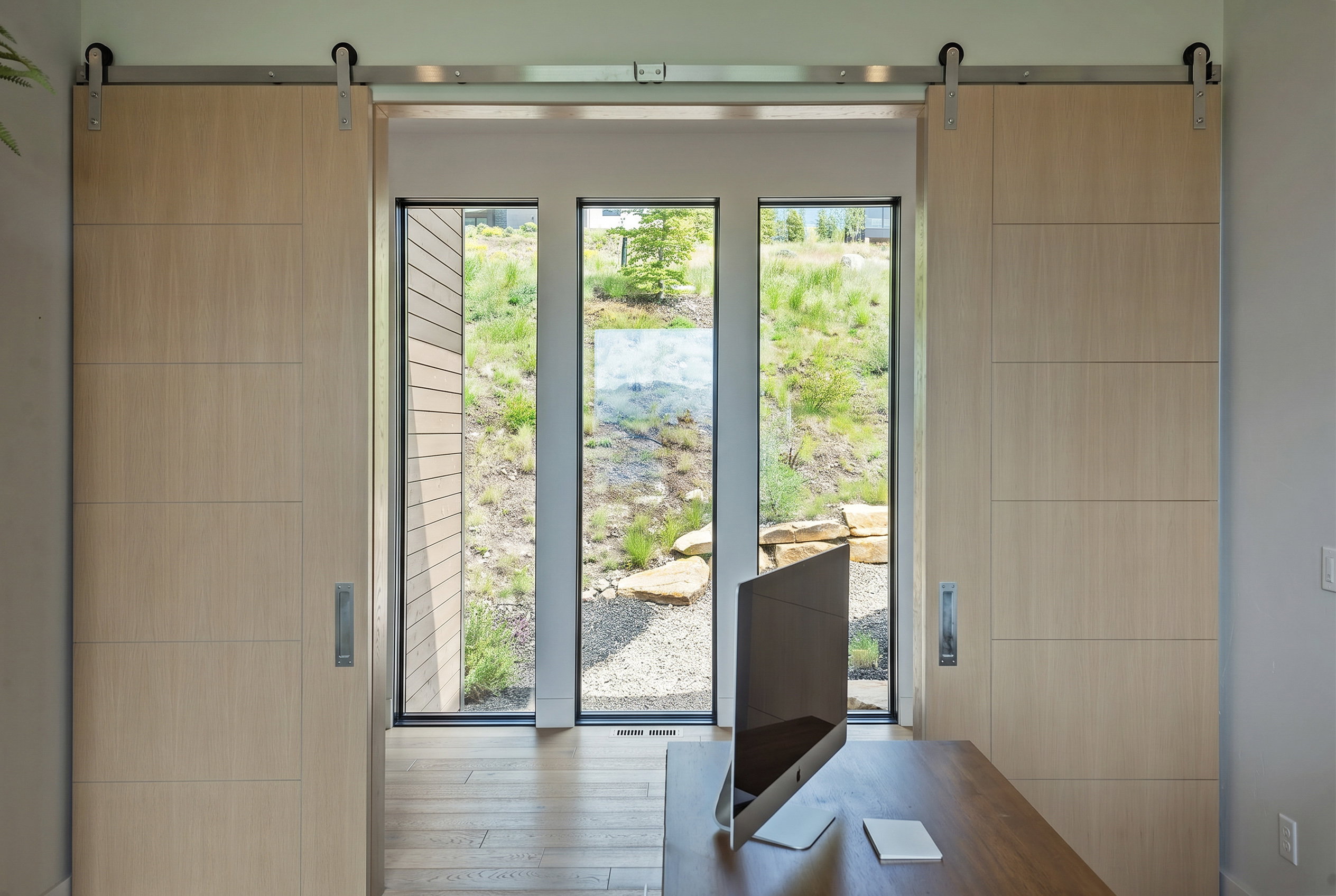 Modern interior space featuring light wood-finish barn doors on a brushed metal track, floor-to-ceiling windows, and wood-finish flooring